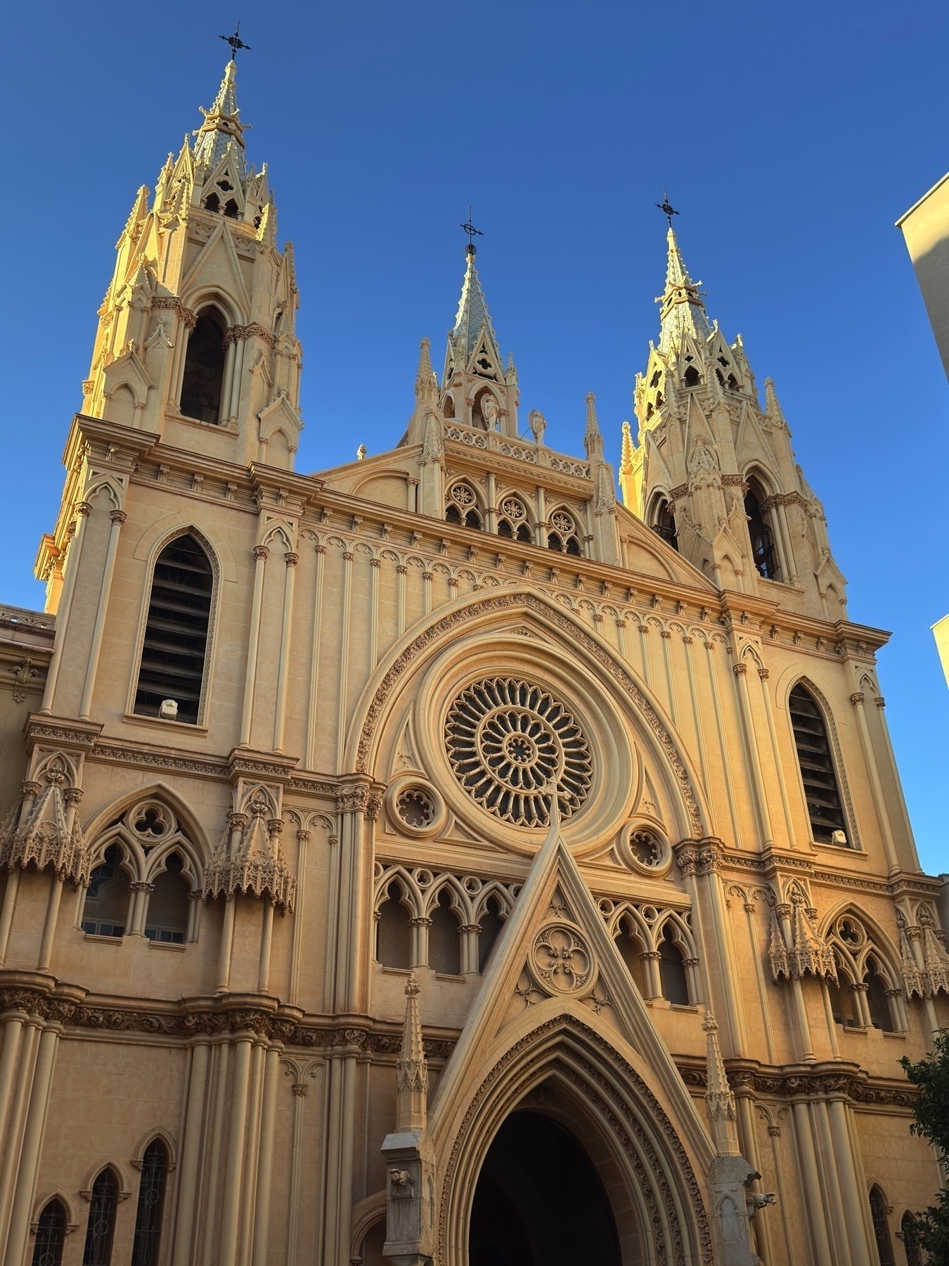 Facade of the Iglesia del Sagrado Corazón in Málaga, featuring three ornate Neo-Gothic spires, pointed arches, pinnacles, and a large rose window above the entrance. Warm light enhances the pale stone against a clear blue sky.