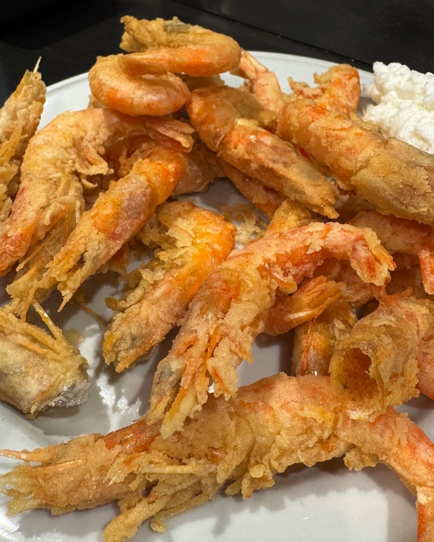 A plate piled with crispy, golden fried shrimp served whole, including heads and tails, highlighting their crunchy texture. A dollop of creamy white sauce sits to the side, adding contrast and a hint of indulgence.
