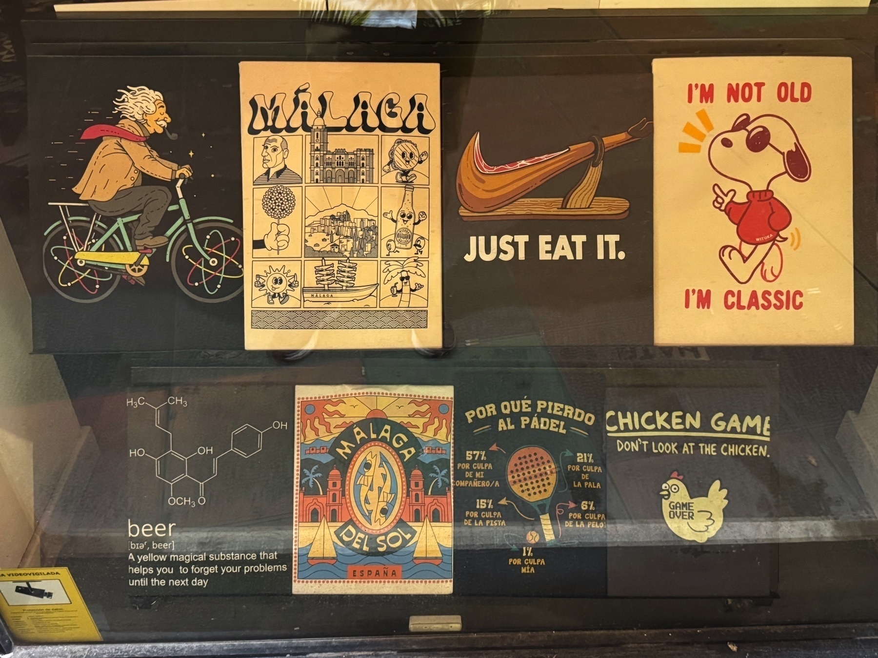 Storefront display of colorful graphic prints on postcards or posters. Designs include: Einstein riding a bike with atoms as wheels; a Málaga city collage; jamón ibérico with “Just eat it”; Snoopy saying “I’m not old, I’m classic”; beer chemical formula; Málaga del Sol design; a paddle tennis joke chart; and “Chicken Game: Don’t look at the chicken” with a cartoon bird labeled “Game Over.”