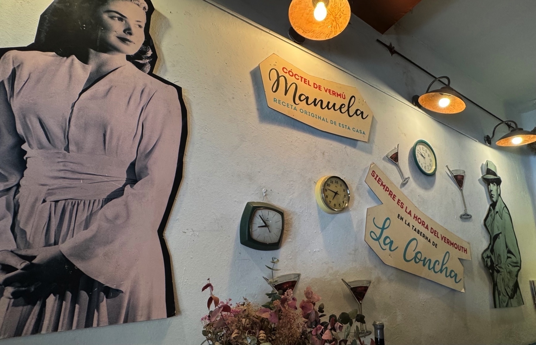 Decorative wall display with life-sized vintage cutouts of a woman and man, mid-century wall clocks, and retro signage promoting the house vermouth cocktail “Manuela.” Martini glass illustrations and warm overhead lighting evoke an old-Hollywood charm with a playful twist.