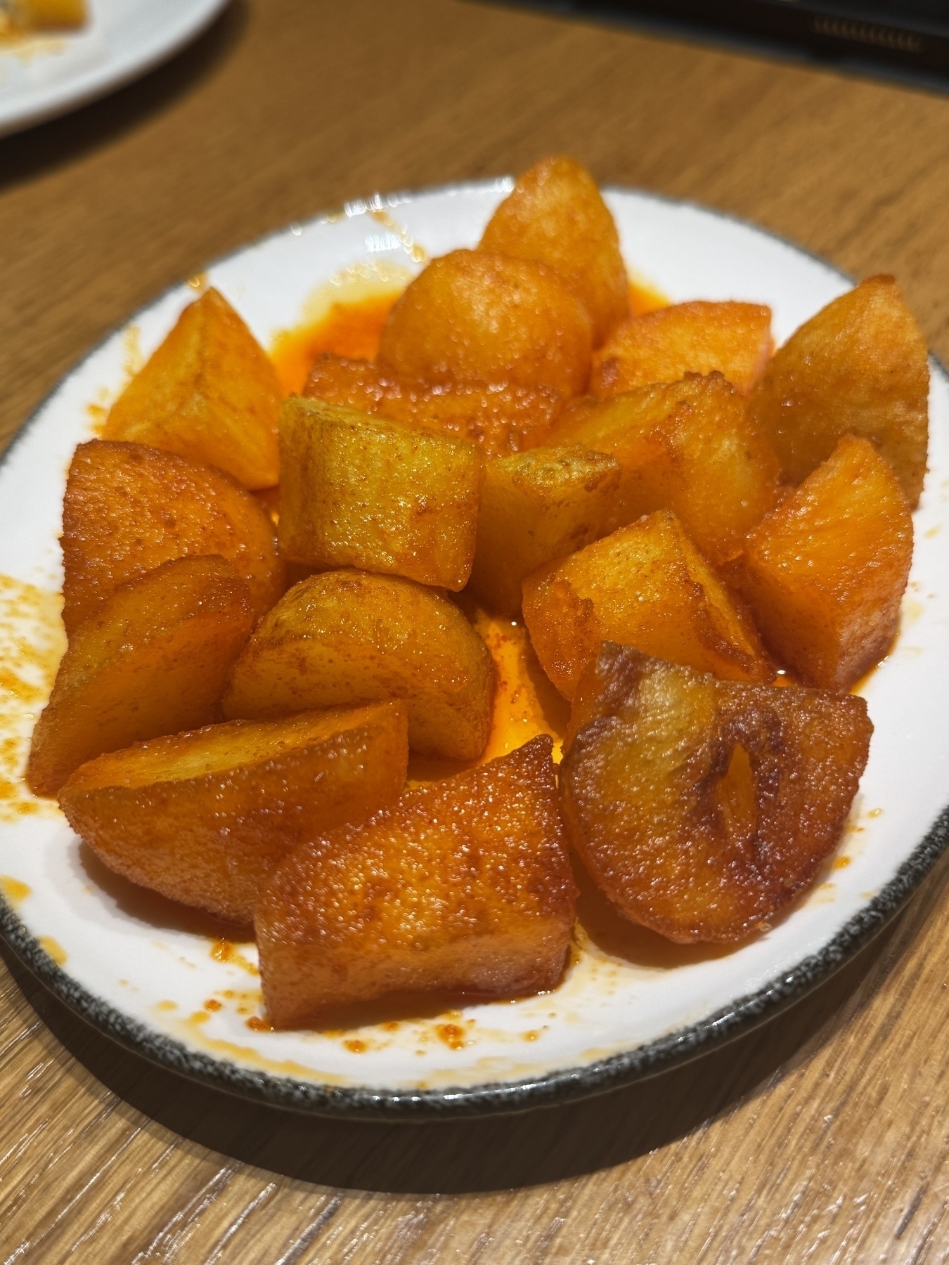 A plate of golden, crispy roasted potatoes in a reddish sauce is placed on a wooden table.