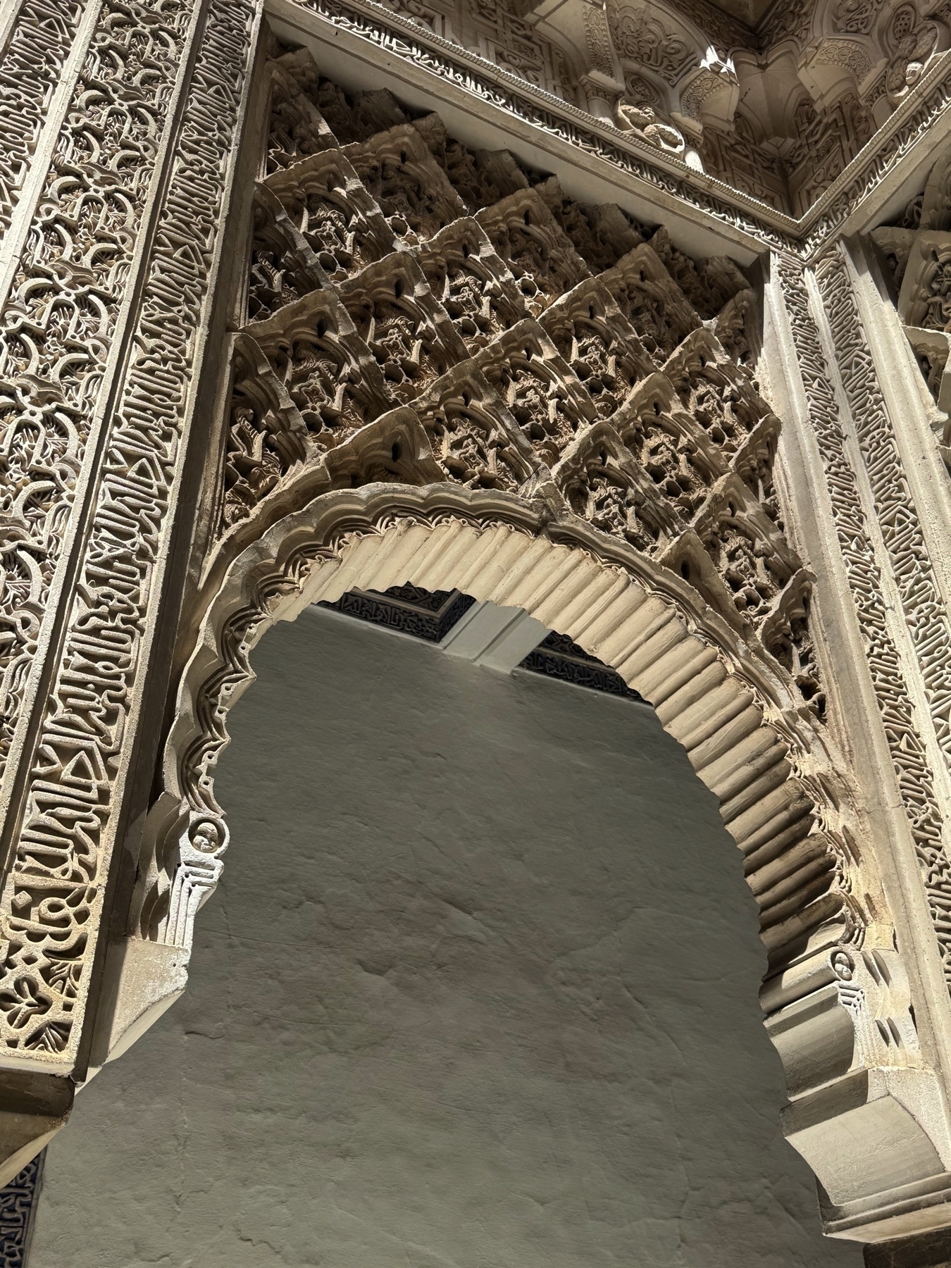 Close-up of a Mudéjar horseshoe arch with intricate plaster detailing, including calligraphic bands, arabesques, and deep geometric latticework. The arch frames a plain white wall beneath a richly decorated spandrel.