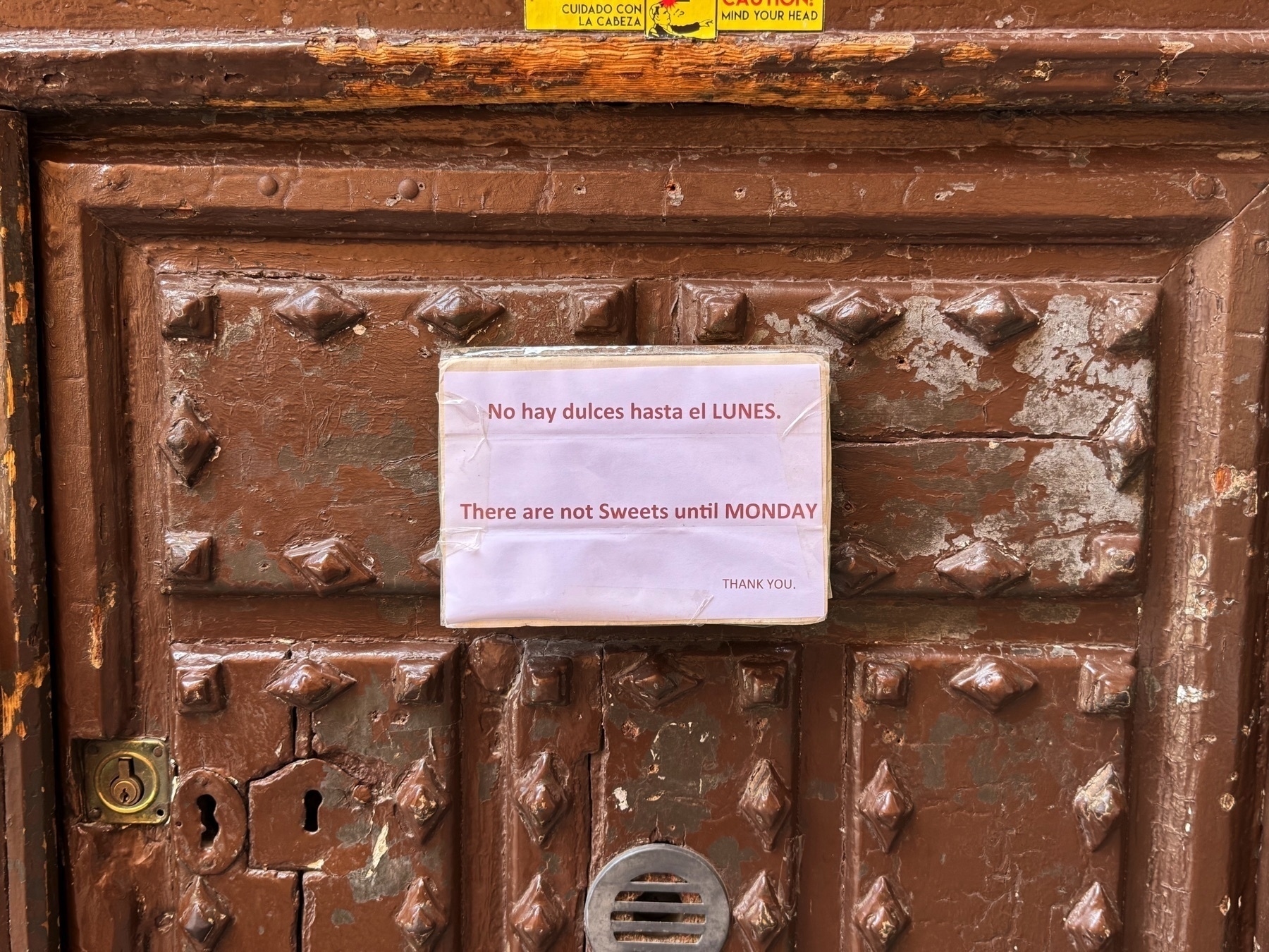 Close-up of a worn brown wooden door with metal studs and a laminated sign taped to the center panel. The sign is printed in both Spanish and English and reads: “No hay dulces hasta el LUNES. There are not Sweets until MONDAY. THANK YOU.” The door’s aged texture and chipped paint add a rustic character to the scene.