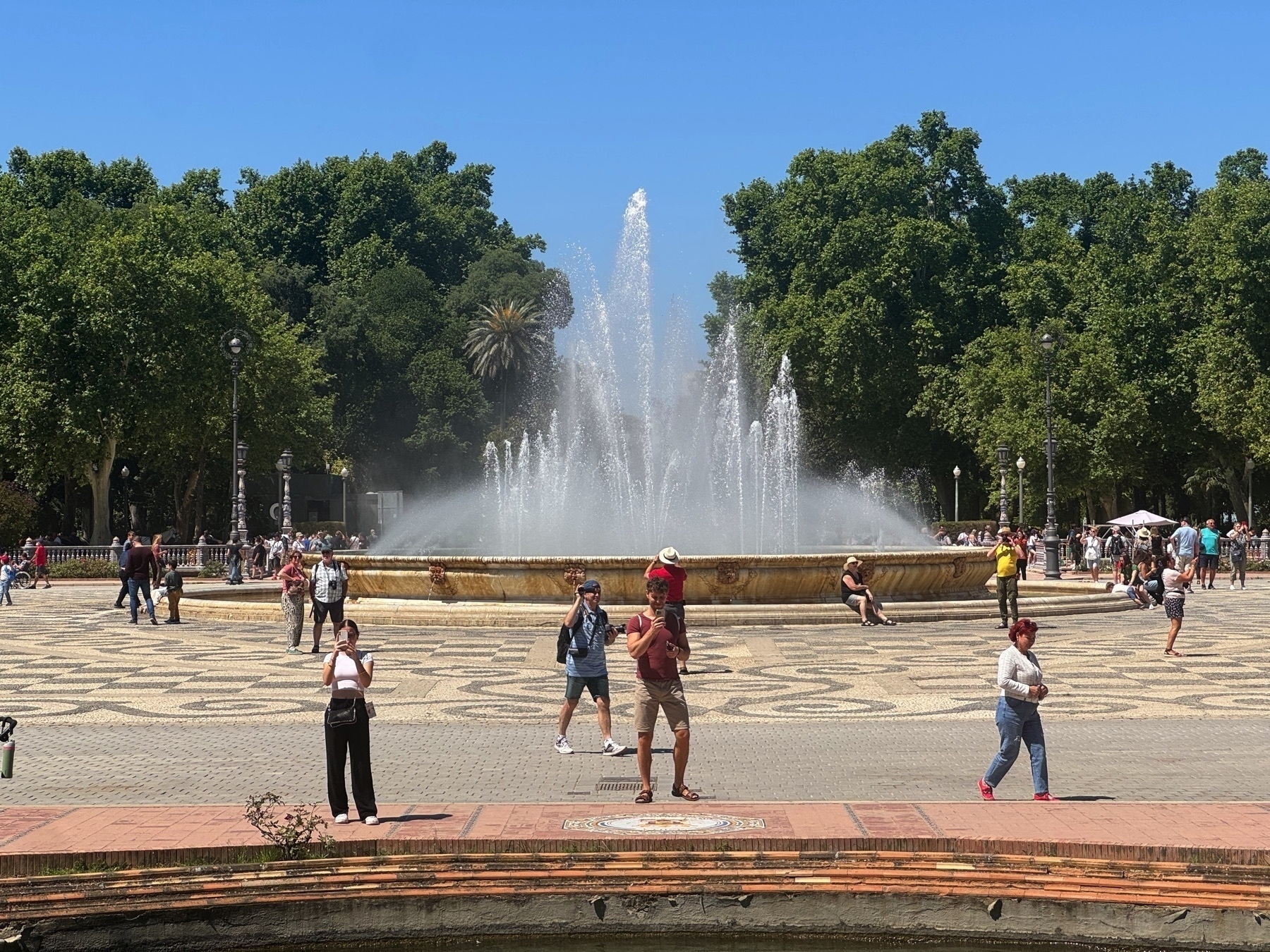 A large circular fountain in Plaza de España sends powerful jets of water into the air, surrounded by a wide, patterned tile plaza where people stroll, sit on the fountain’s edge, and take photos. A man in shorts taking a photo on his phone feels like a mirror to the photographer. The backdrop features dense green trees, ornate lampposts, and a bright blue sky, evoking a lively, summery atmosphere.