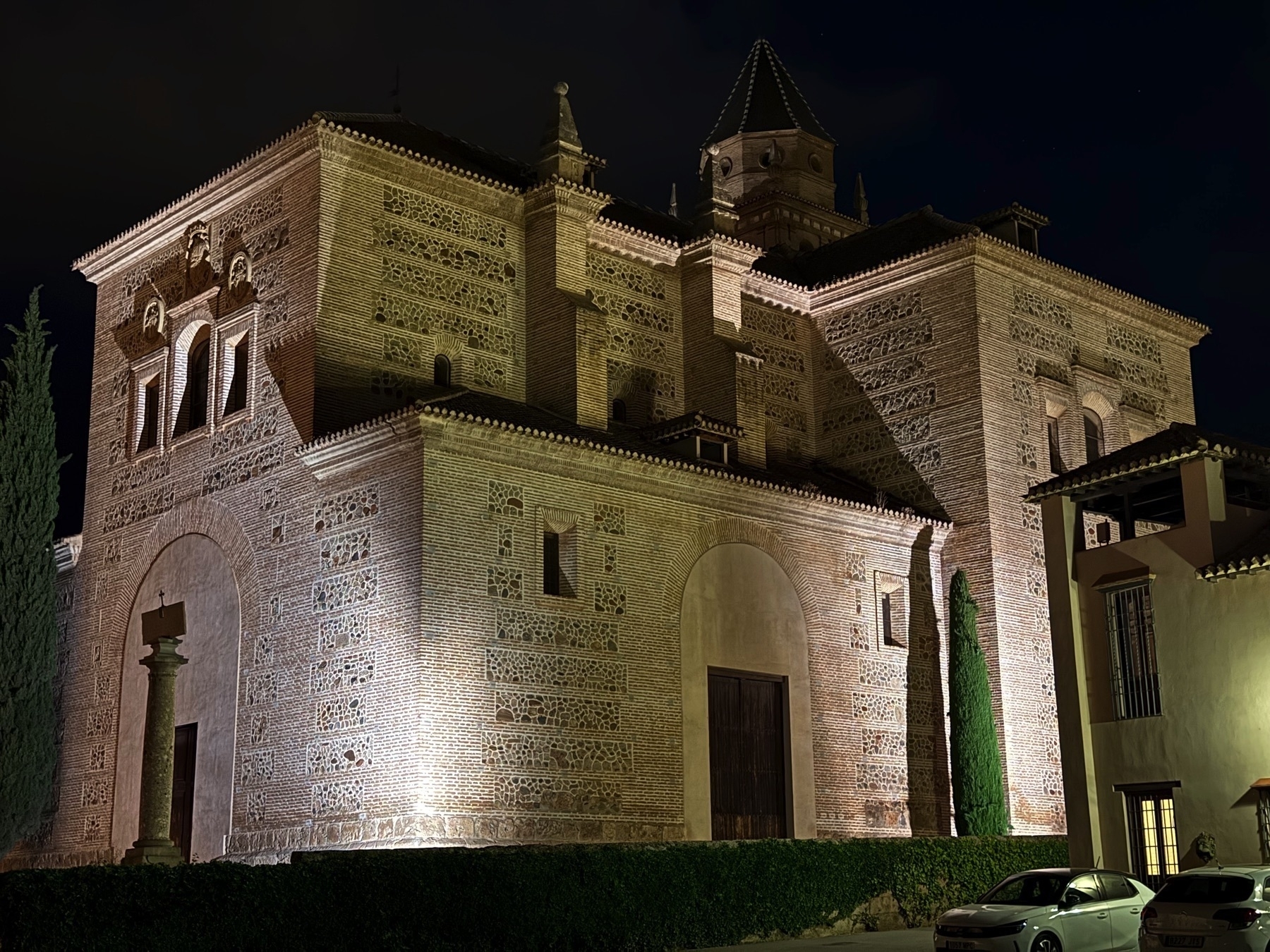 Night view of the Church of Santa María de la Alhambra, its richly textured brickwork and Renaissance details highlighted by exterior lighting, with cypress trees and nearby structures in shadow.