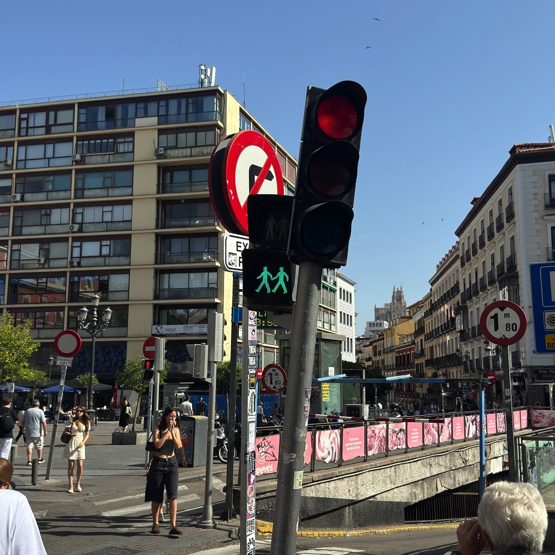 Street scene in Madrid showing a pedestrian traffic light with a green signal featuring two figures holding hands, commonly used as a symbol of LGBTQ+ inclusion. The surrounding area includes modern and historic buildings, various street signs, and several pedestrians walking or waiting.