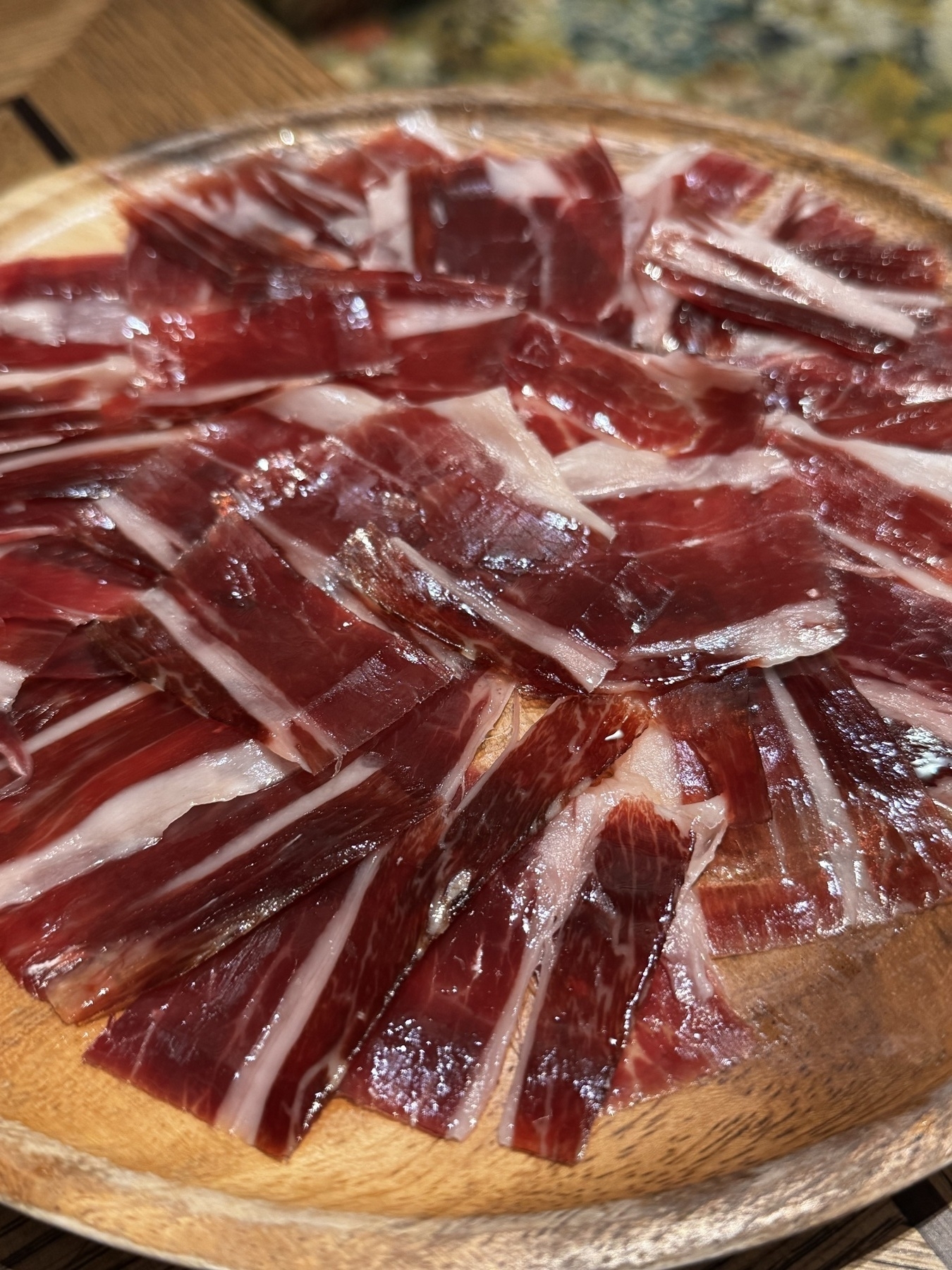 Thin slices of glossy, marbled jamón ibérico arranged in a fanned, overlapping pattern on a round wooden plate.