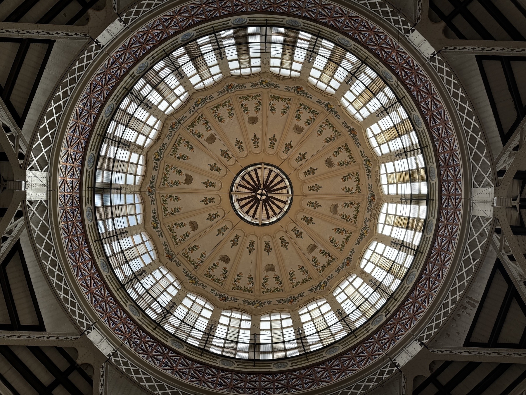 Interior view of the central dome of Mercat Central de València, featuring an ornate circular skylight with floral frescoes and radiating decorative patterns, framed by metal latticework and large arched windows allowing natural light.