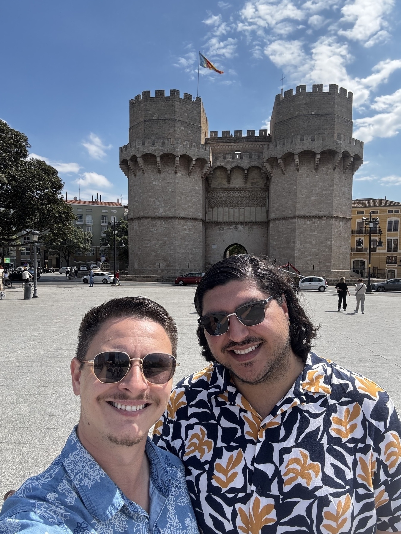 Chasen and his partner pose for a selfie in front of the imposing Torres de Serranos, a medieval stone gate with crenellated twin towers and a fluttering flag overhead. The bright sun and open plaza give the scene a joyful, vacation-like feel.