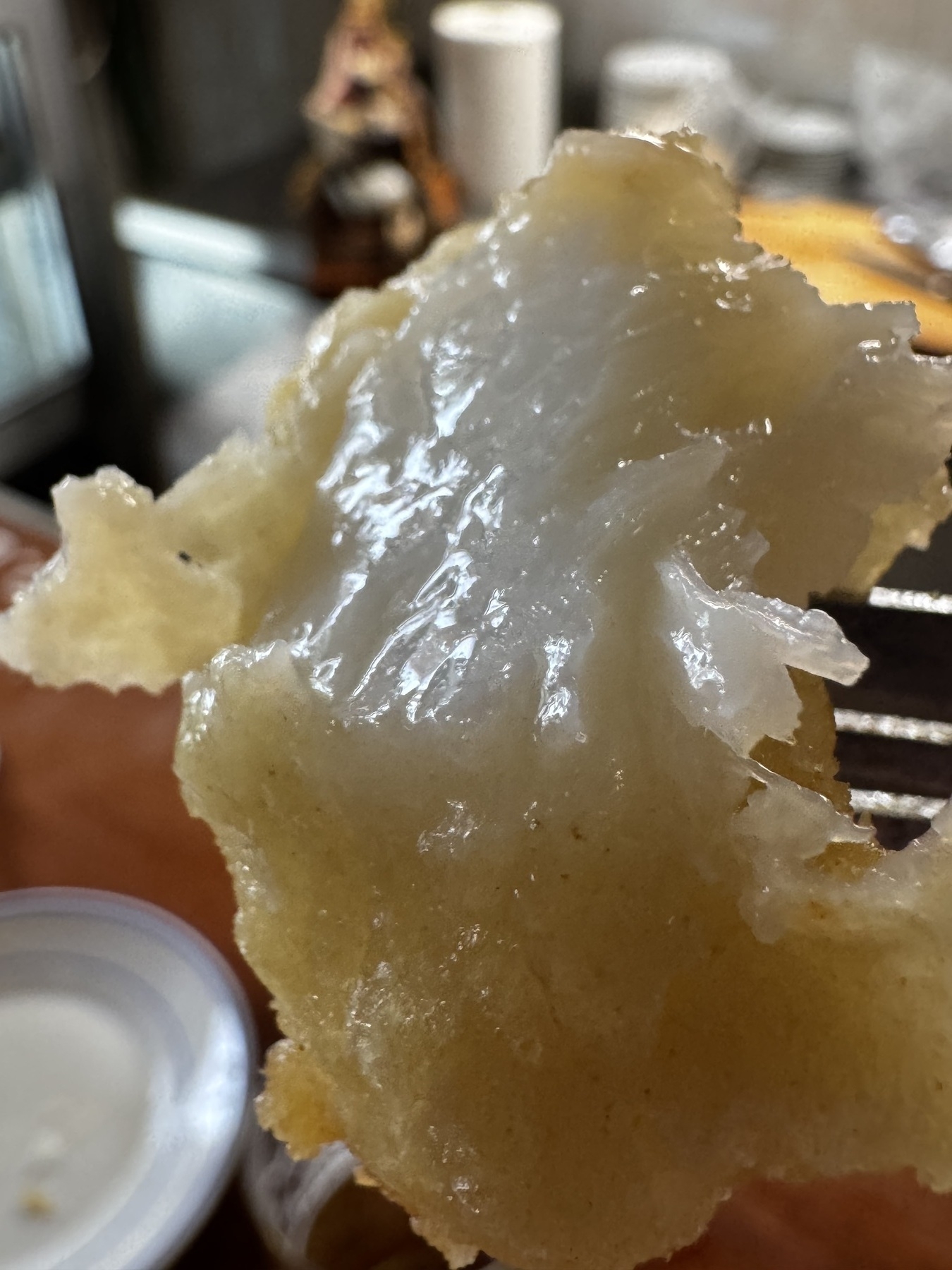 Close-up of a flaky, translucent piece of fried cod with a golden, crispy outer edge, held up against a blurred kitchen background.