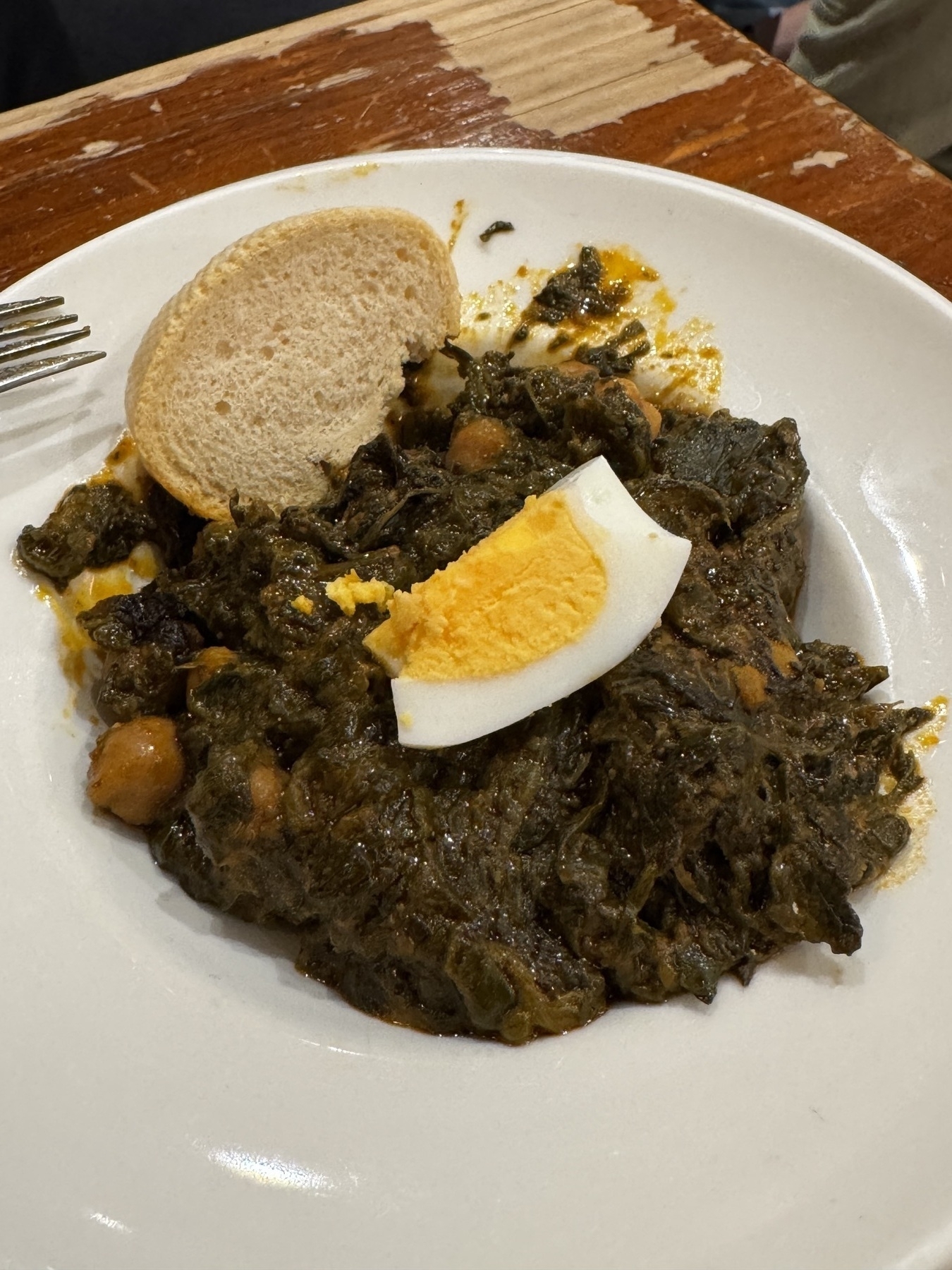 A white plate of espinacas con garbanzos, a traditional Andalusian stew of sautéed spinach and chickpeas in a dark, savory sauce. The dish is topped with a wedge of hard-boiled egg and served with a slice of crusty white bread. The hearty texture and deep color make it look rustic and nourishing.