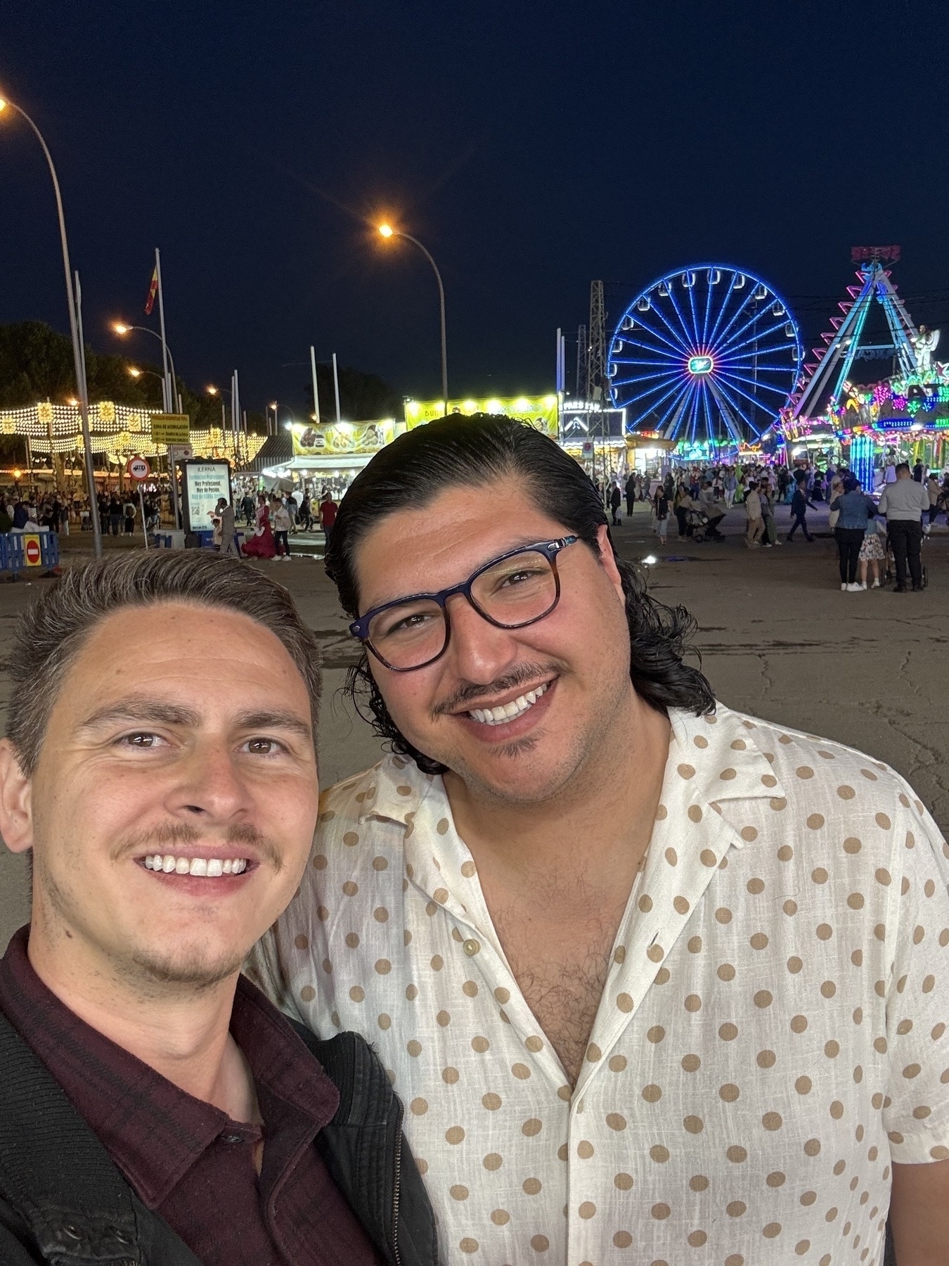 Two men smiling and posing for a selfie at night with a large Ferris wheel, thrill rides, and food stands illuminated in colorful neon lights behind them. The festive atmosphere and bright backdrop capture the energy of the event’s final night.