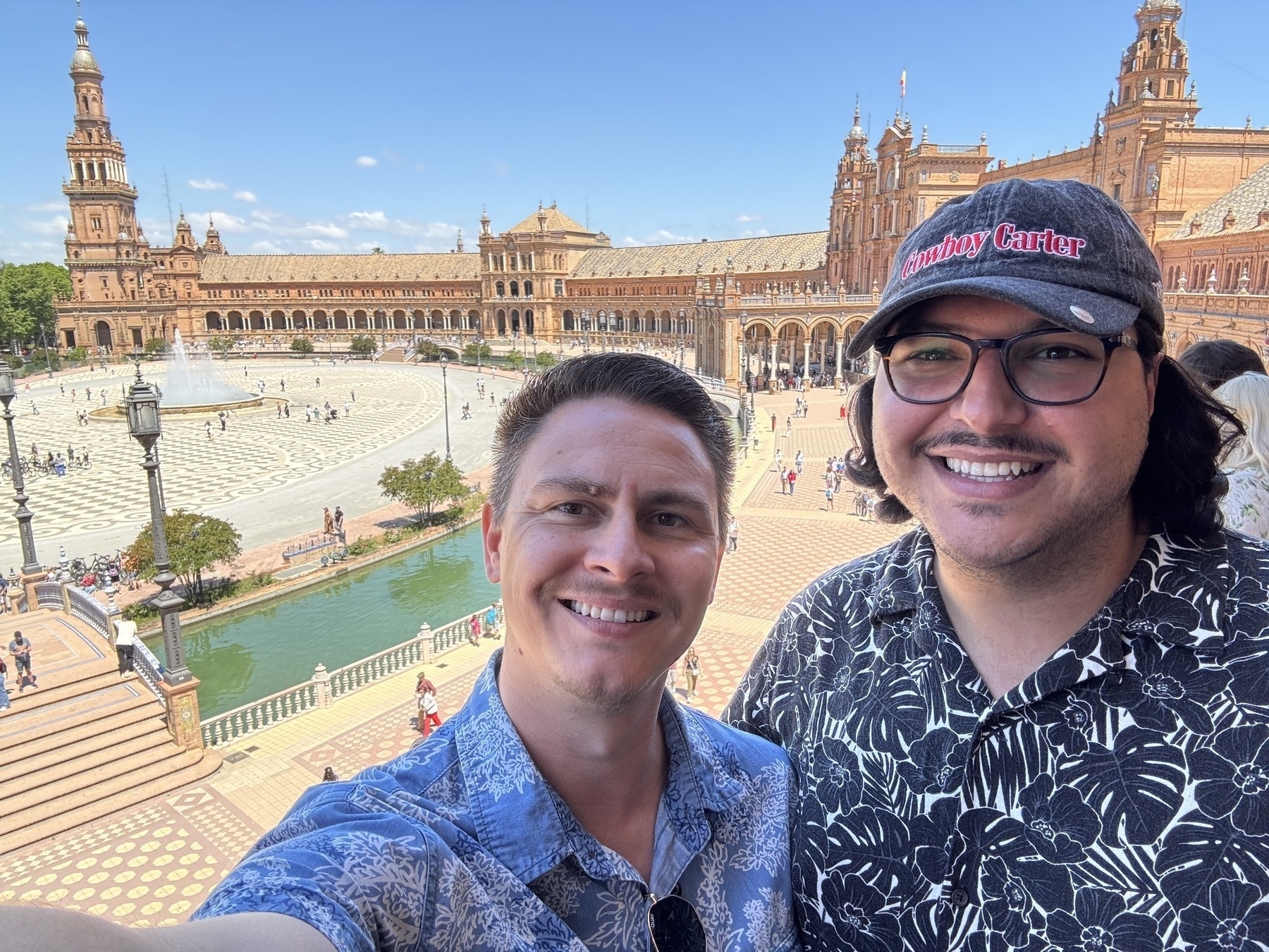 Two smiling men pose for a selfie on a sunny day from an elevated viewpoint. Behind them is the Plaza de España’s grand semicircular building with arched colonnades, a central tower, tiled walkways, a canal with green water, and a central fountain surrounded by visitors.