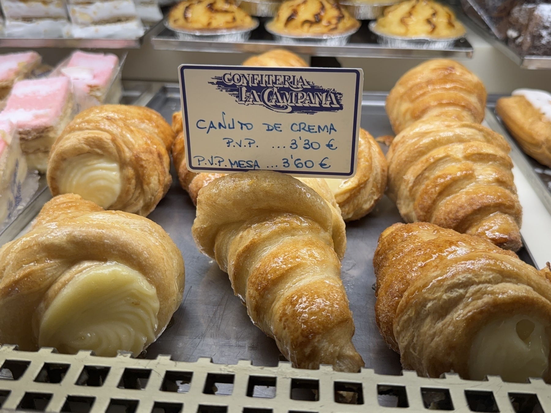 Close-up of a display case at Confitería La Campana showing rows of glossy, golden-brown puff pastries labeled “Canuto de Crema.” Each pastry is swirled into a horn shape and filled with rich, pale custard cream that peeks out from the open ends. The pastries look flaky and buttery, with a light sheen that suggests a sweet glaze, making them appear freshly baked and indulgent.