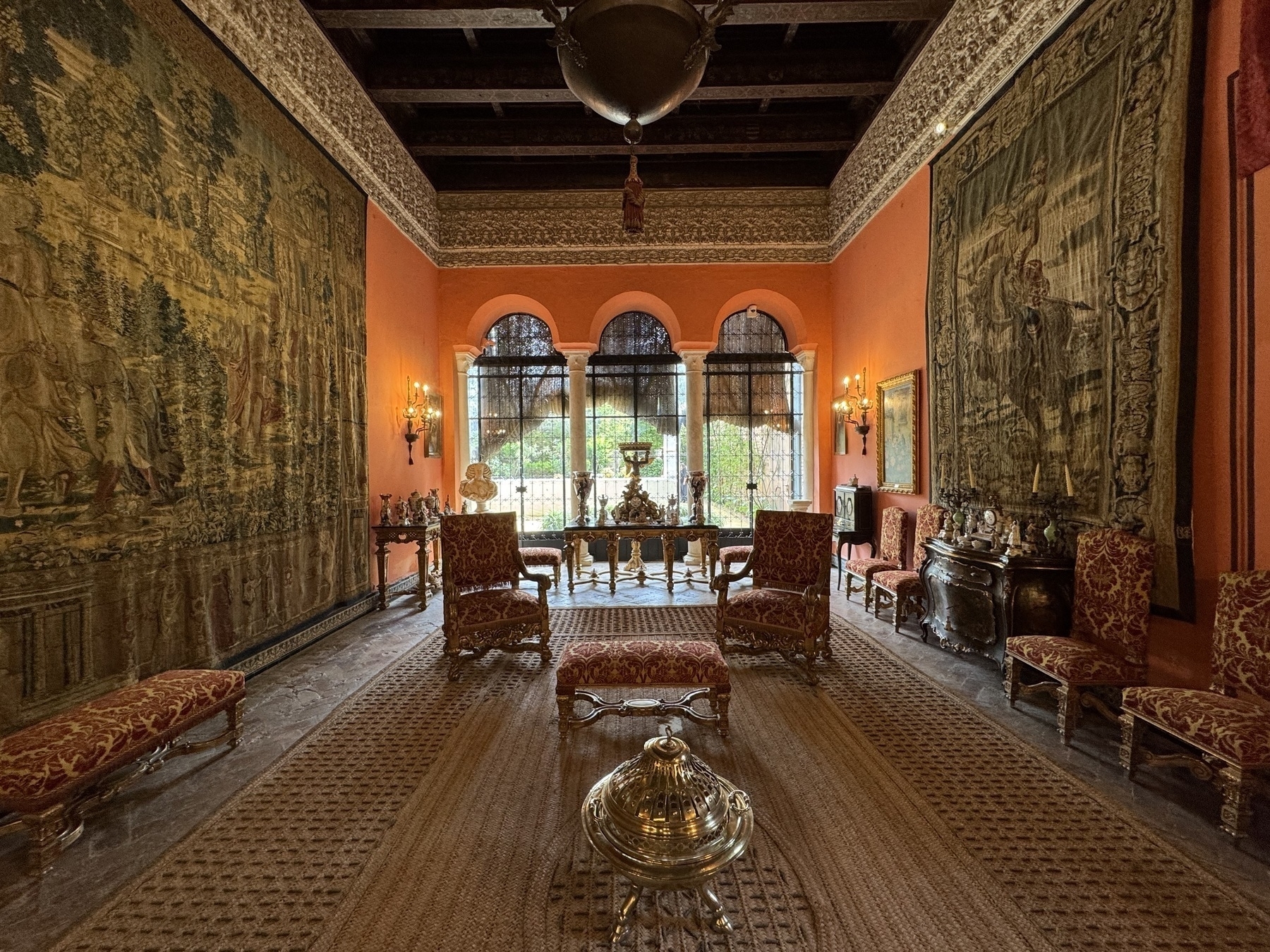 An ornate sitting room with dark wooden beams and orange walls, decorated with large medieval-style tapestries, gilded furniture, and a collection of antique objects arranged on side tables.