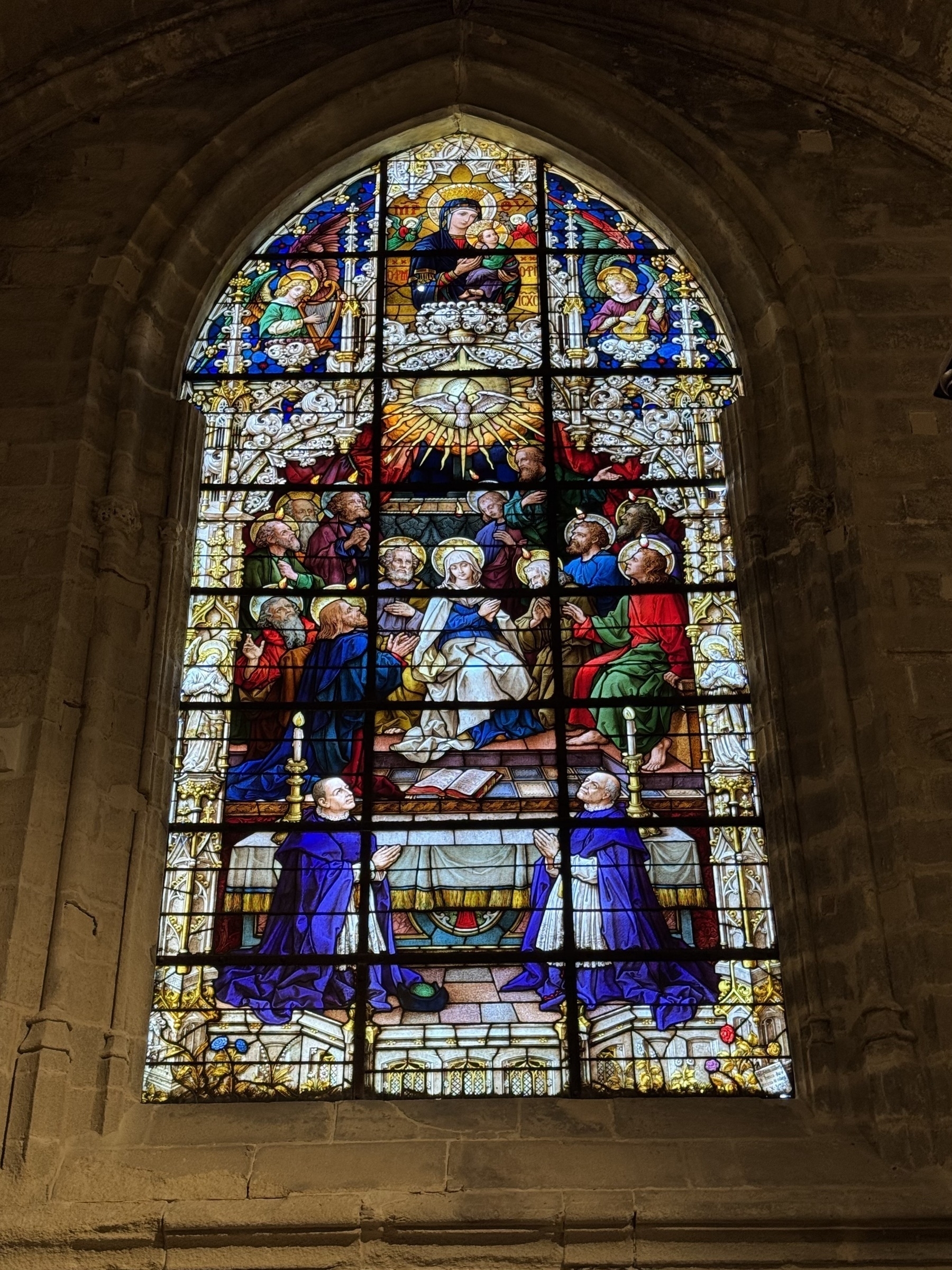 Richly colored stained glass window depicting a religious scene with the Virgin Mary and child surrounded by apostles, angels, and kneeling clergy, framed by pointed Gothic stonework.