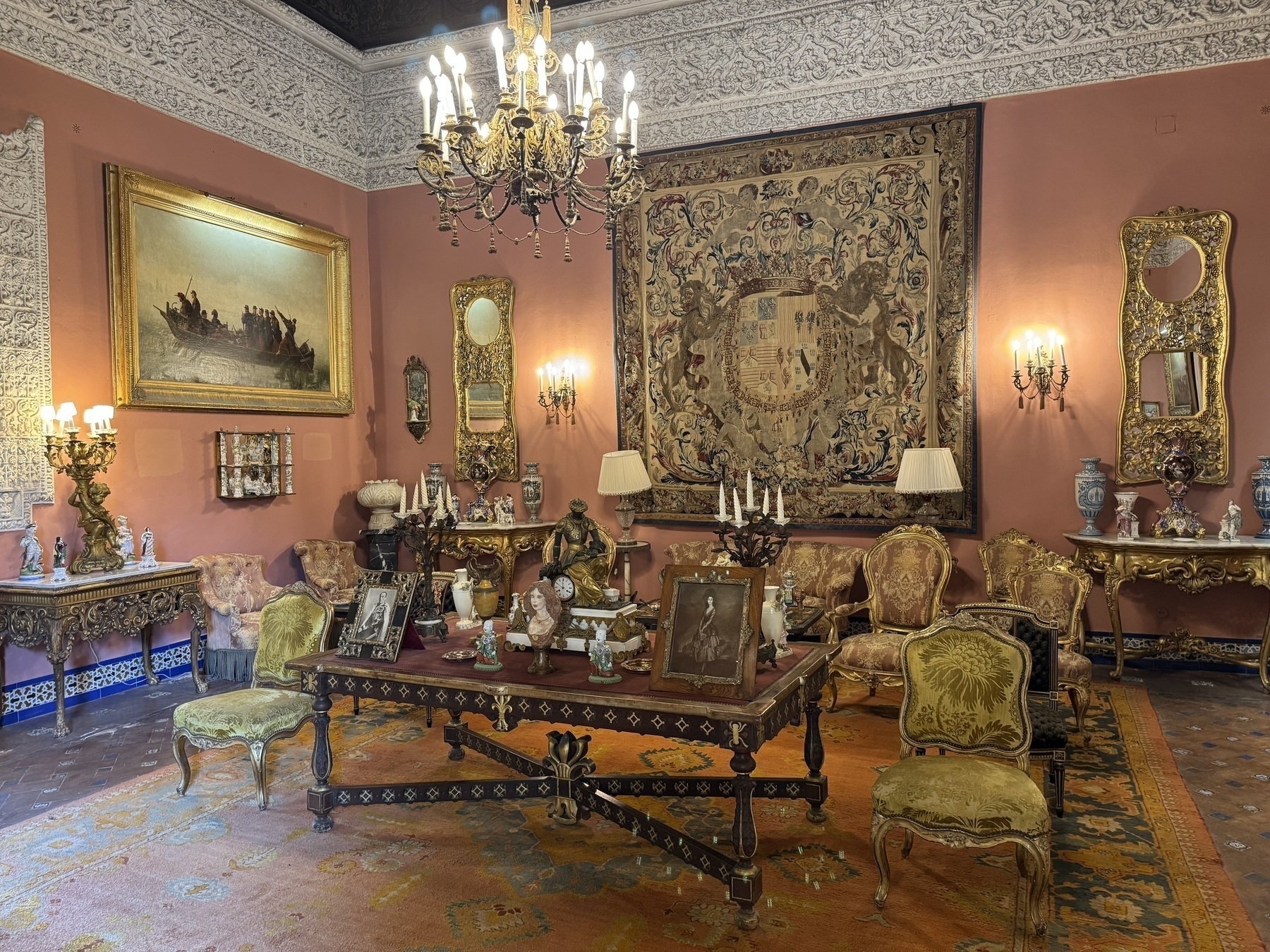 A richly decorated salon featuring a large coat-of-arms tapestry, antique furnishings, ornate mirrors, and a grand chandelier, with porcelain vases and framed artwork adding to the lavish decor.