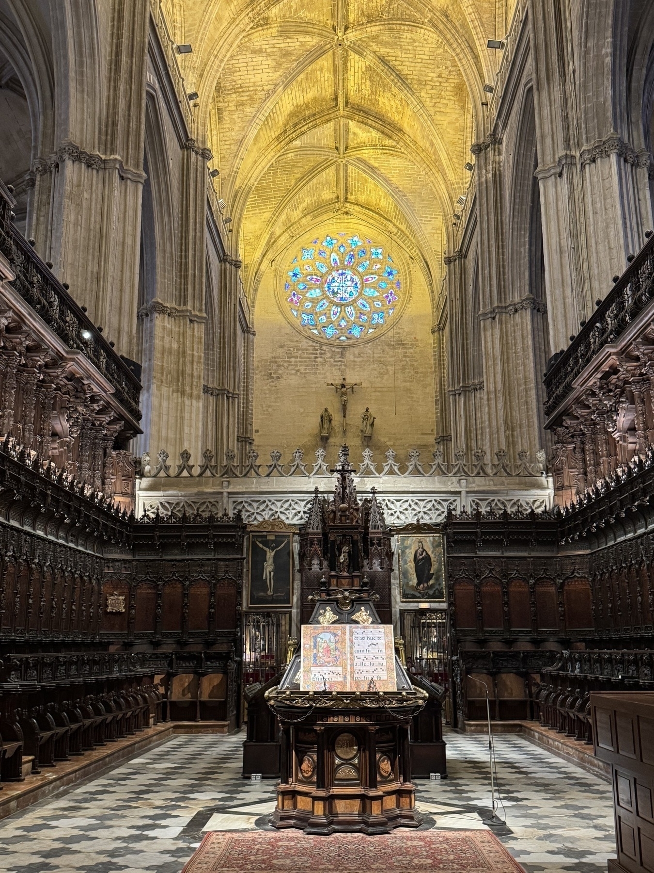 Interior view of the cathedral’s elaborately carved wooden choir stalls with high vaulted ceilings above, a brilliant rose window glowing in the apse, and a central lectern displaying illuminated manuscripts on a richly patterned rug.