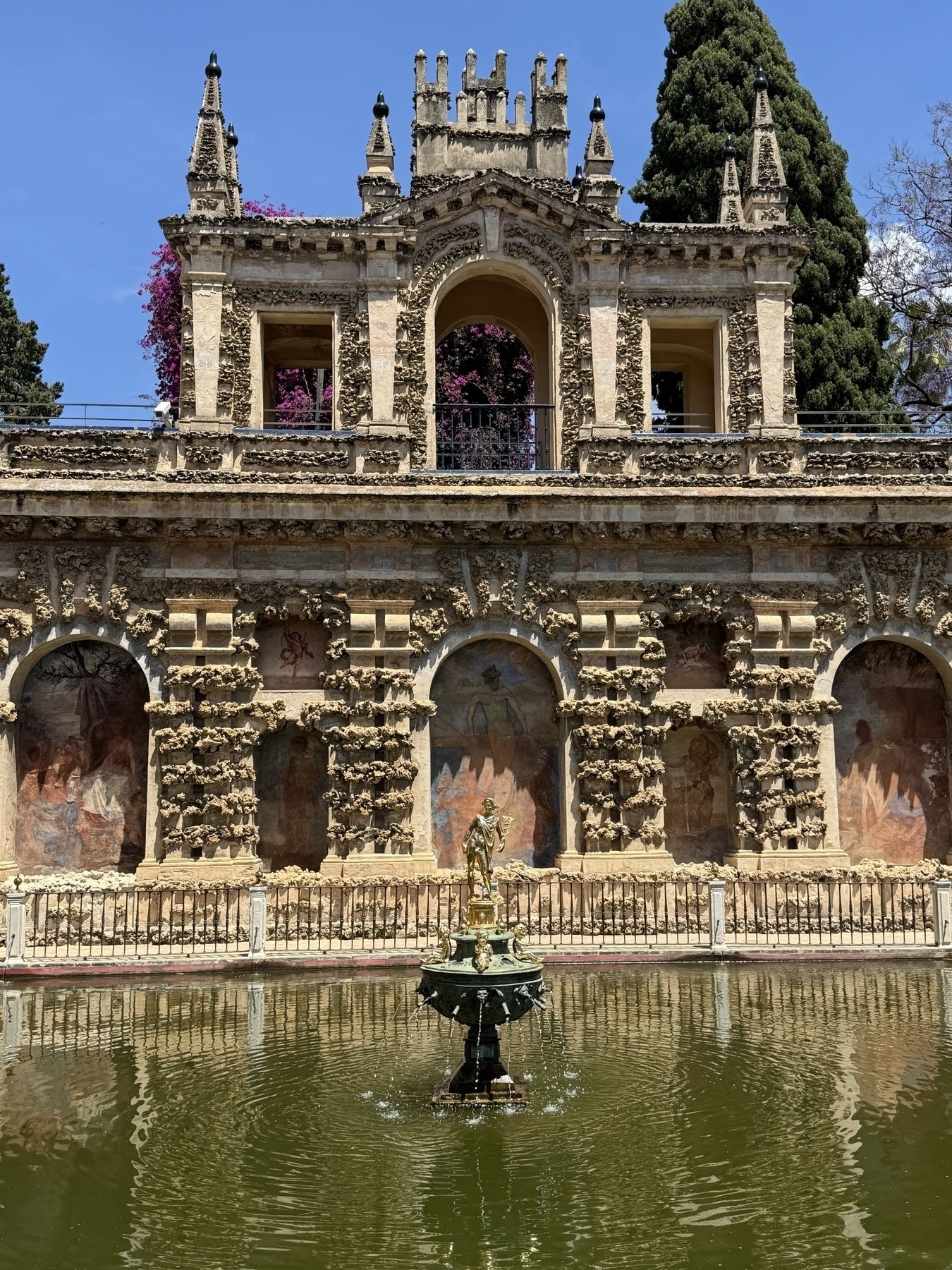 The Gallery of the Grotesque and Mercury Pond, showing a Renaissance arcade with rustic stucco niches, faded frescoes, and an upper gallery framed by pointed pinnacles. At the center of the pond stands a bronze statue of Mercury on a decorative fountain base.
