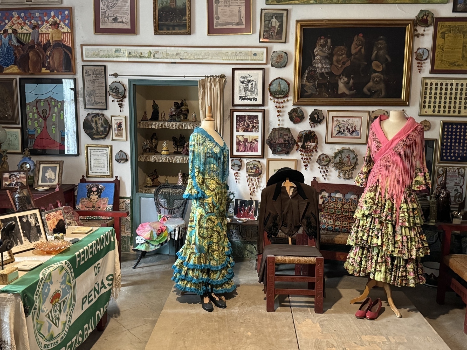 A museum-like room filled with traditional Andalusian memorabilia, including two mannequins in colorful flamenco dresses, a short jacket on a chair, and walls densely covered with framed posters, paintings, and decorative plates.
