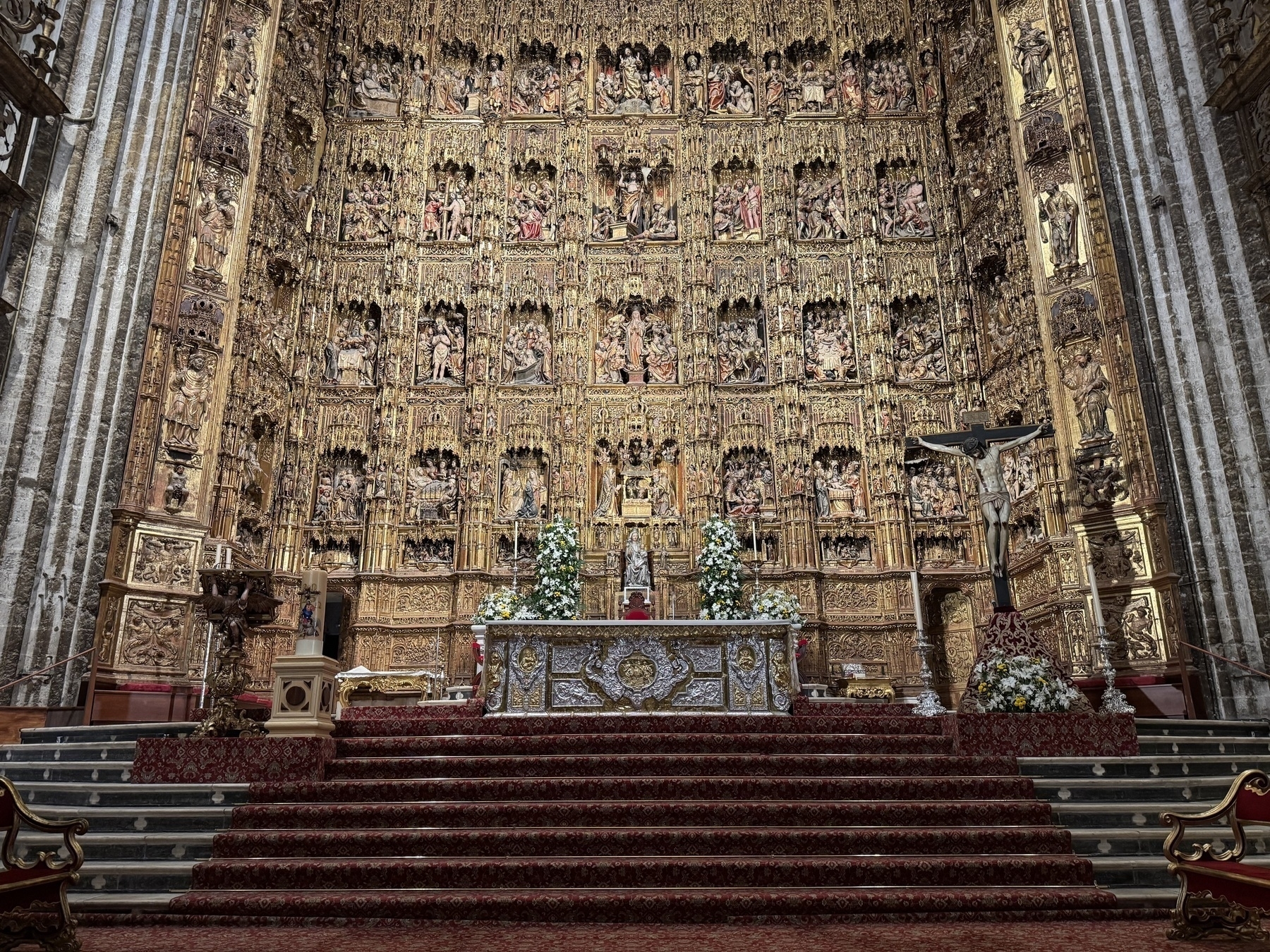 Stunning view of the cathedral’s main altar, fronted by ornate steps and flanked by floral arrangements, with a towering gilded altarpiece behind it—densely packed with intricate carvings depicting scenes from the life of Christ and other biblical figures.