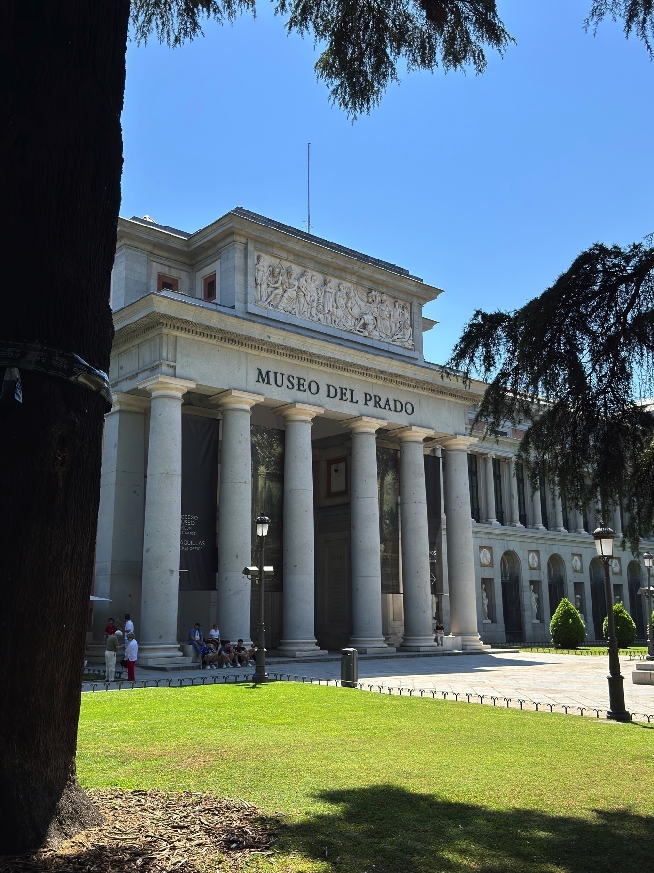 Angled view of the Museo del Prado’s entrance framed by trees, emphasizing the symmetry of the neoclassical architecture and the texture of the stone columns. A small group of people rests in the shade beneath the columns. The sky is bright and clear, and the surrounding greenery adds contrast to the pale stone facade.