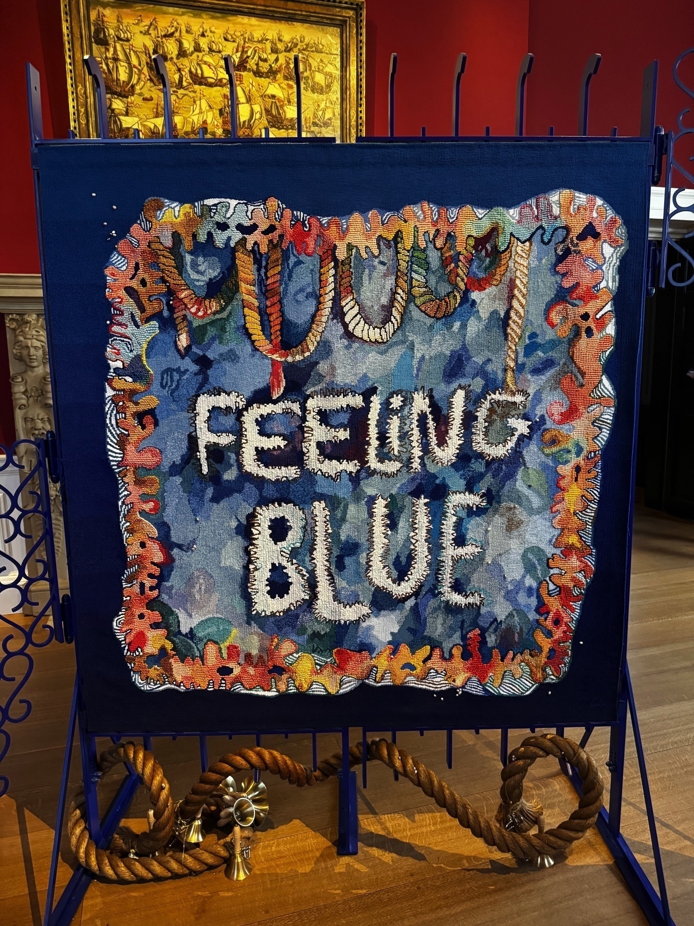 A contemporary textile piece displayed in a gallery space with deep red walls and wooden flooring. The artwork reads “FEELING BLUE” in stitched white letters on a mottled blue background, bordered by colorful embroidered patterns resembling sea creatures and coral. Nautical rope and decorative metal elements at the base add a tactile, whimsical touch to the installation.