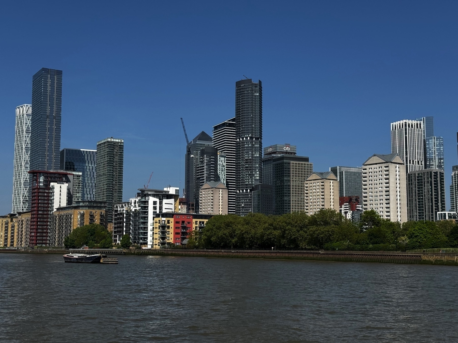 A skyline view of Canary Wharf, with a dense cluster of high-rise buildings featuring varied modern architectural styles. The Thames flows calmly in the foreground, and a low line of trees separates the river from the cityscape.