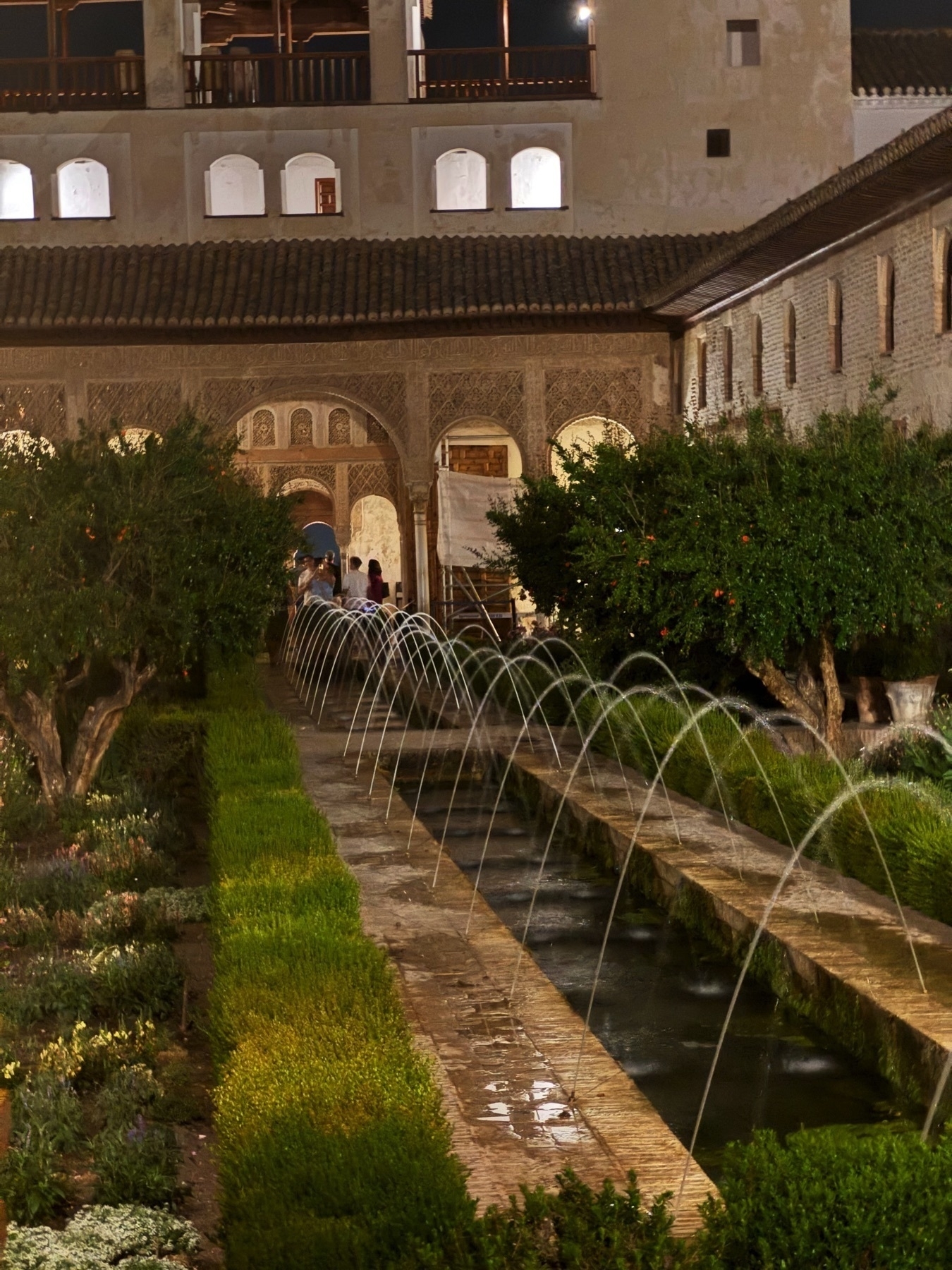 The Generalife’s Patio de la Acequia at night, with a central water channel bordered by arching fountains and symmetrical garden beds, leading toward a colonnaded hall adorned with intricate stucco work.