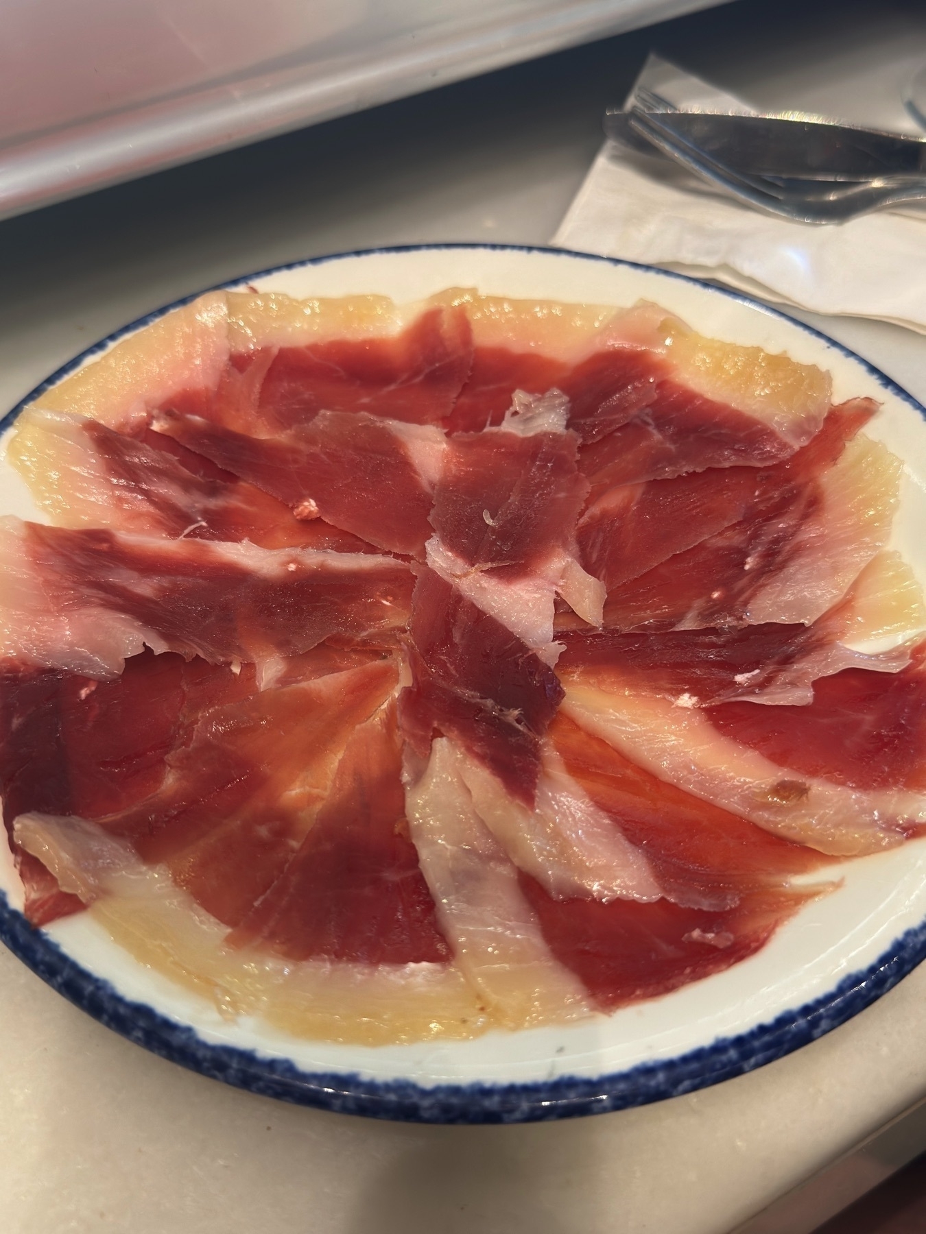 A circular arrangement of thinly sliced jamón ibérico fanned out across a white plate with a blue rim. The deep reds and glossy fat edges on the ham shimmer under the light, making it look tender and luxurious.