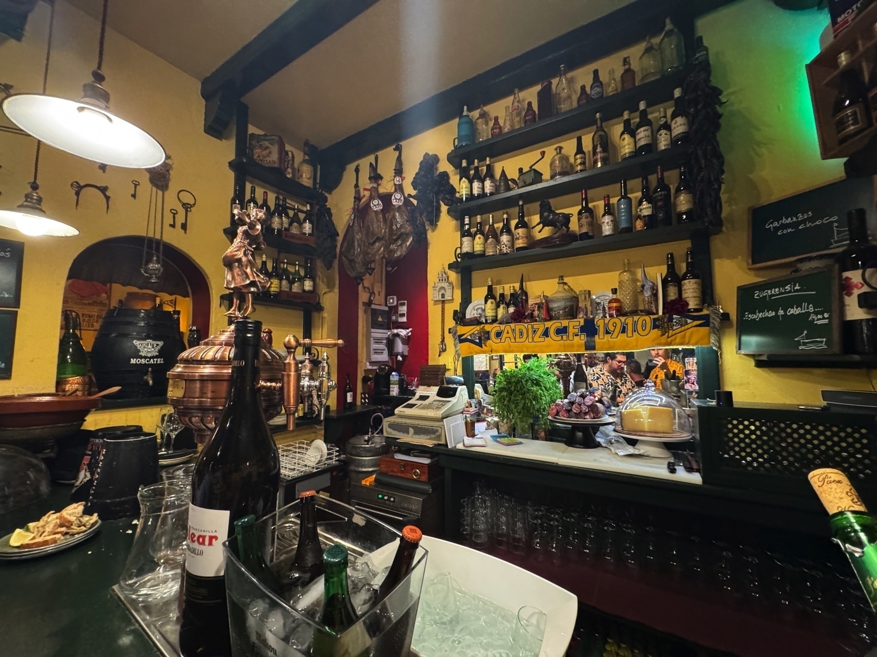 The interior of a traditional Cádiz-style taberna, warmly lit and filled with character. Hanging cured hams, shelves of vintage bottles, and rustic wooden beams frame the scene. A Cádiz C.F. 1910 scarf draped across a mirror adds local pride, while fresh herbs, grapes, and cheeses at the bar create a cozy and authentic atmosphere.