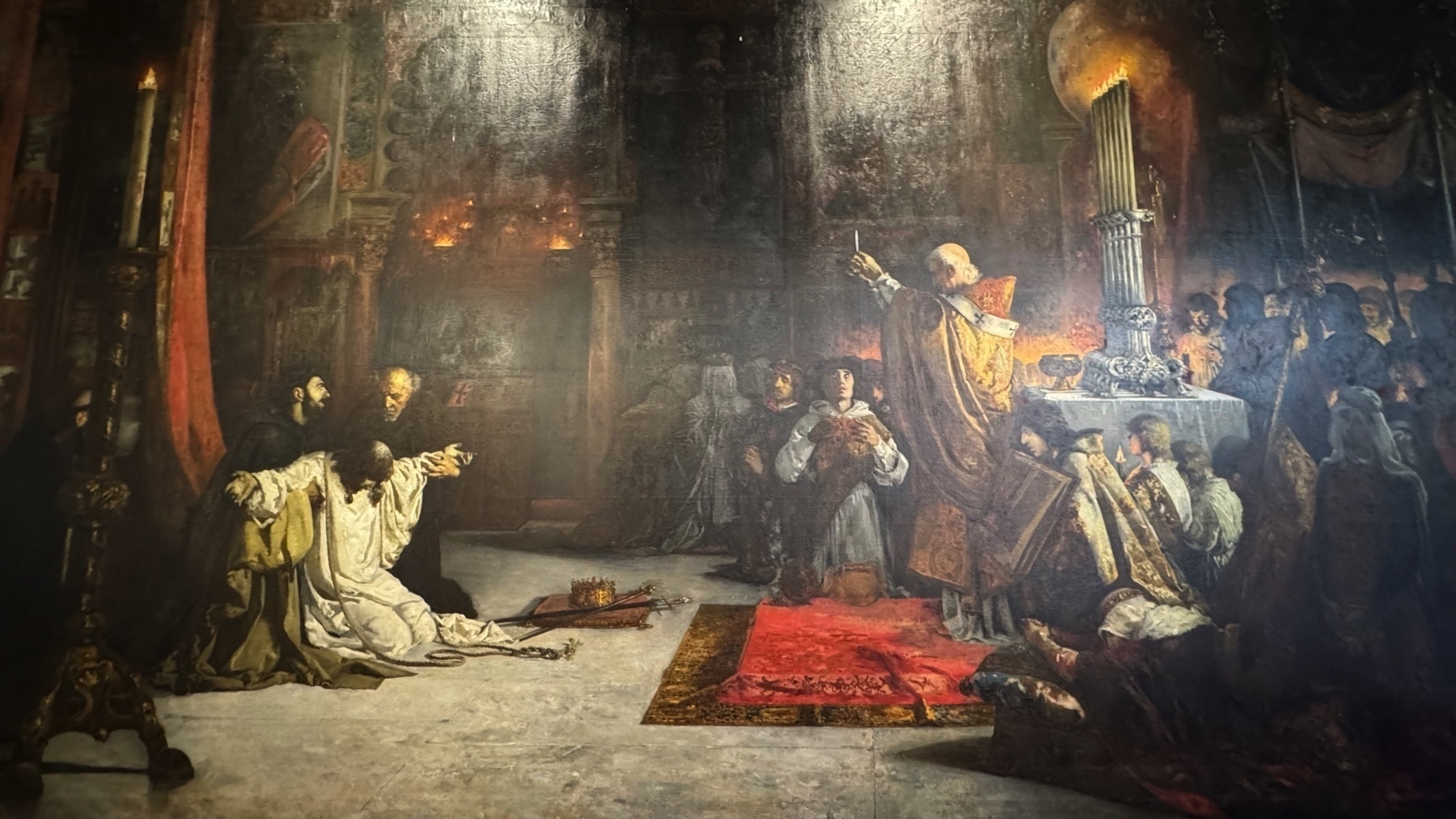 A painting inside the palace depicting a dramatic religious scene, with a kneeling figure held by monks and a bishop celebrating Mass. The composition includes richly textured garments, candlelight, and architectural backdrops rendered in a dark, Baroque style.