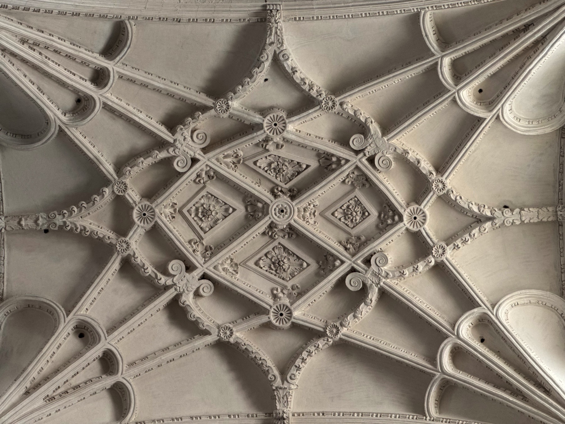 Vaulted ceiling detail with a symmetrical ribbed pattern, featuring floral and scroll motifs carved in white stone. The design showcases the intricacy of Gothic vault construction, with intersecting arches and decorative bosses creating a harmonious, geometric composition that draws the eye toward the center.