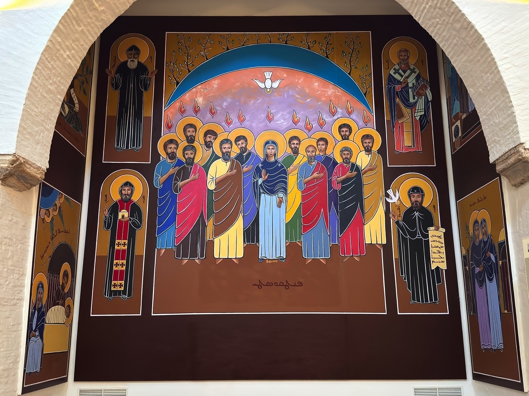 Religious mural showing a group of saints and apostles surrounding the Virgin Mary, with flames of Pentecost above their heads and a white dove at the top symbolizing the Holy Spirit. Bold lines and bright, flat colors define the stylized iconography.