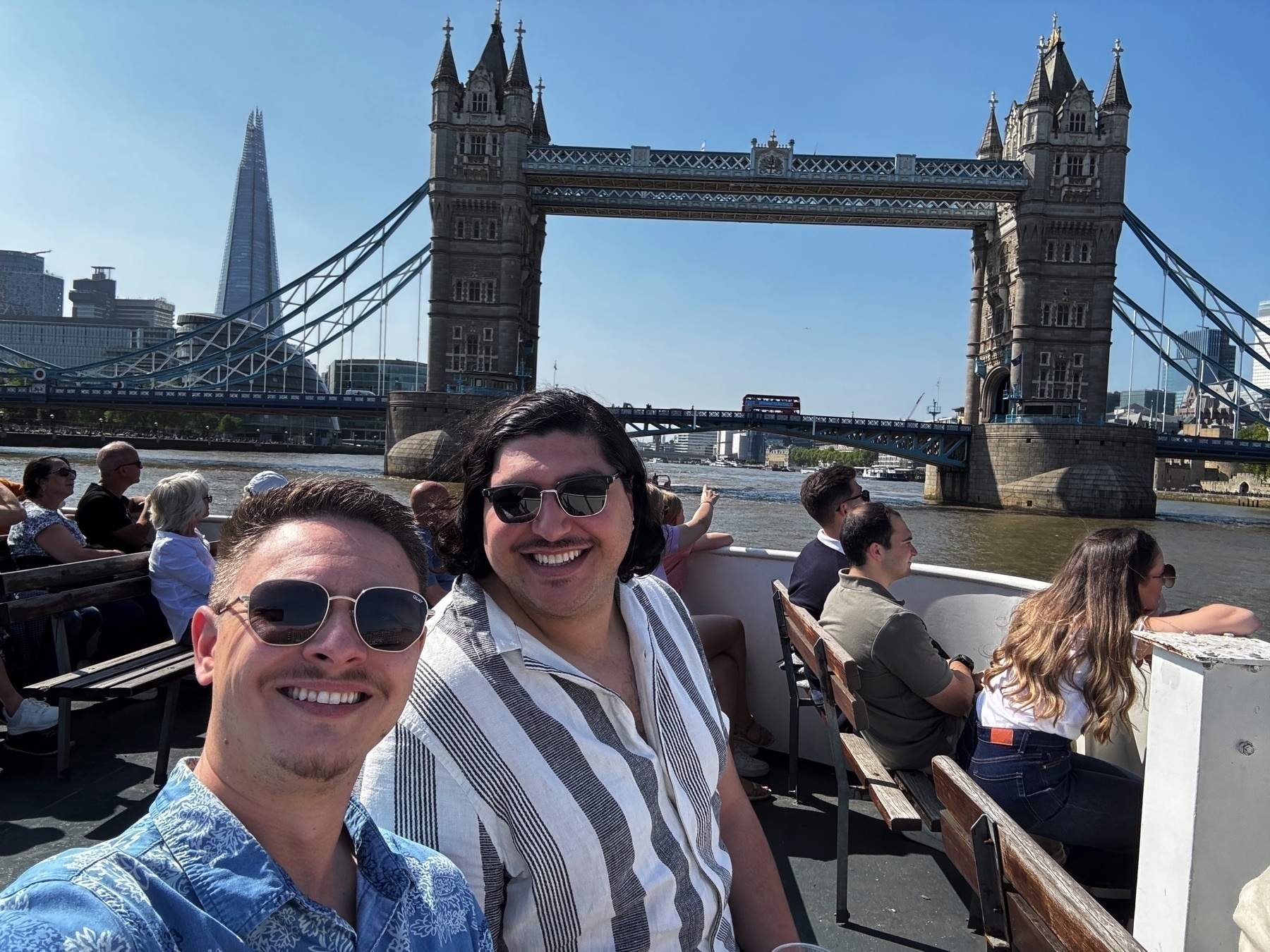 Two smiling men sit on the deck of a boat with Tower Bridge prominently in the background, flanked by the Shard and modern London skyscrapers. Other passengers are seated behind them, enjoying the sunny weather and Thames River views.