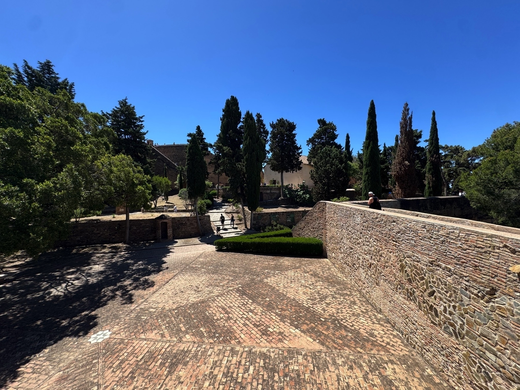A peaceful courtyard surrounded by stone walls and cypress trees, with patterned brick walkways and trimmed greenery. Several people walk or rest under the clear blue sky, adding a sense of scale and serenity to the historical setting.