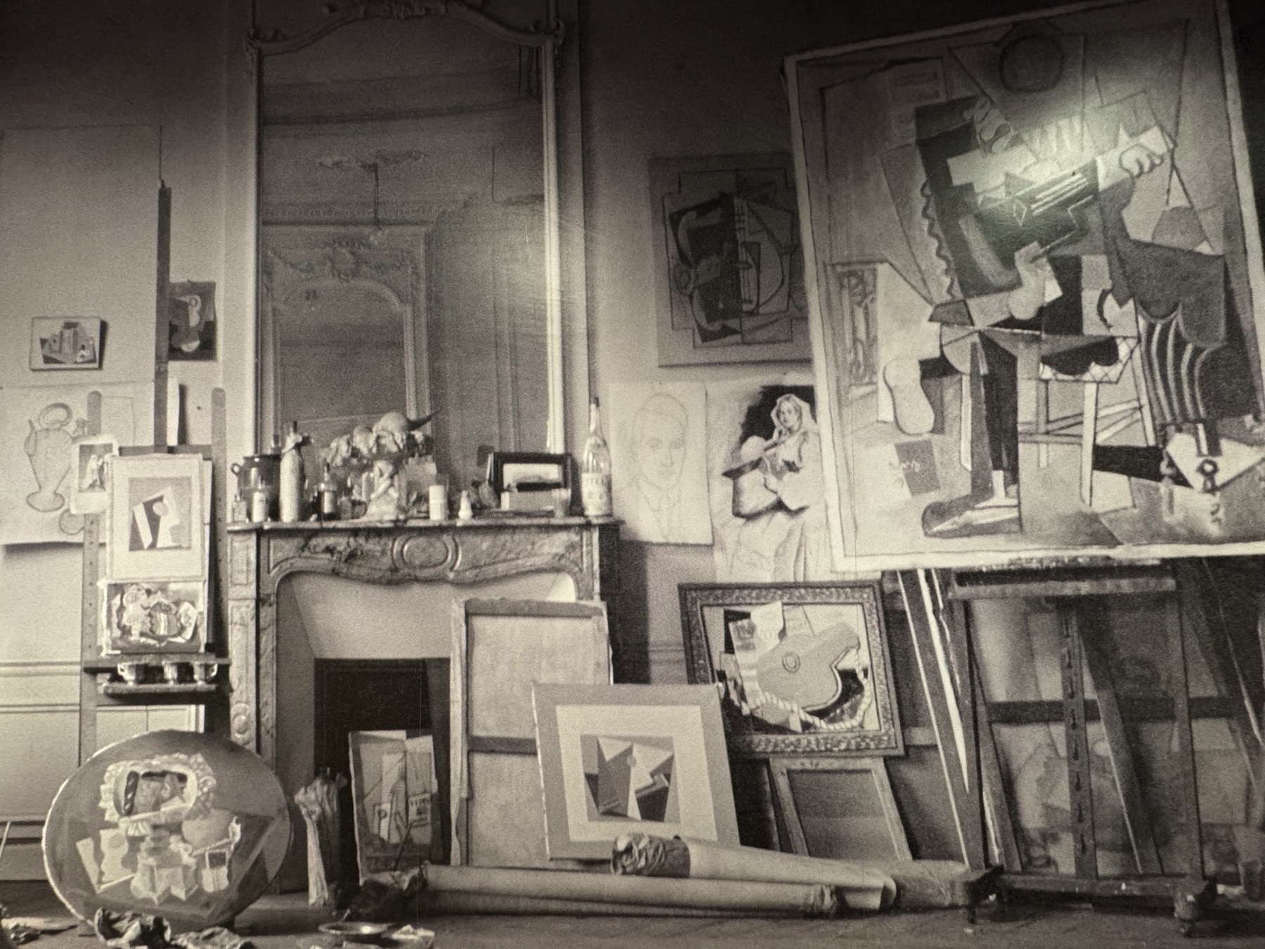 A black-and-white photograph of Pablo Picasso’s studio at Rue Schoelcher, Paris, showing numerous Cubist artworks in various stages of completion. A large canvas prominently displays a fragmented figure playing a guitar, likely from Picasso’s Synthetic Cubism period, where bold lines and overlapping planes build abstracted forms. The space is filled with paintings, drawings, and sculptural props, highlighting the artist’s intense productivity and experimental process in early 20th-century modernism.