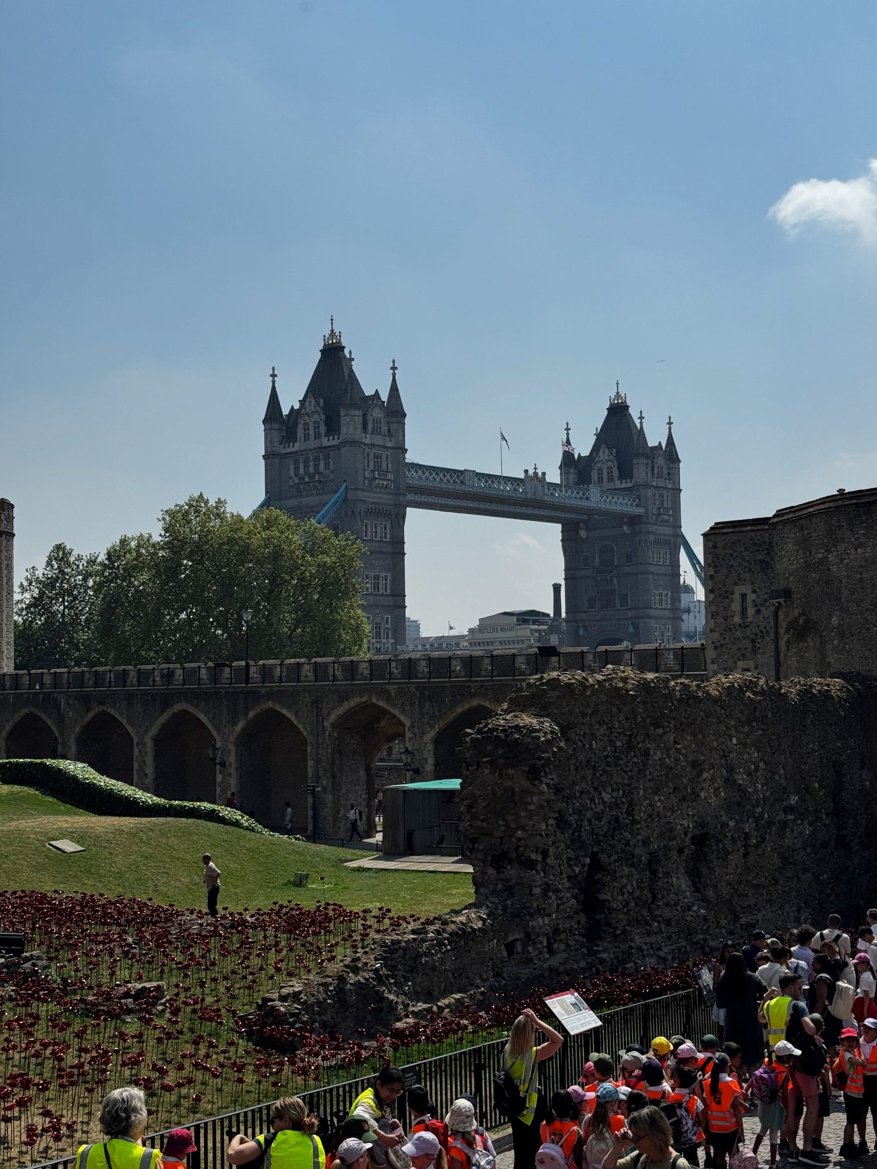 Tower Bridge’s twin towers frame the background beyond arched stone walkways and grassy lawns, with remnants of old walls and a crowd of visitors in bright clothing adding depth and lively scale to the historic scene.