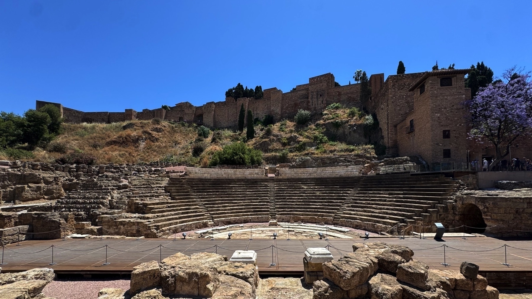 Panoramic view of the Roman Theatre in Malaga with tiered stone seating and a wooden stage area below. Behind the theatre, the fortified walls and towers of the Alcazaba rise along the hillside under a cloudless blue sky.