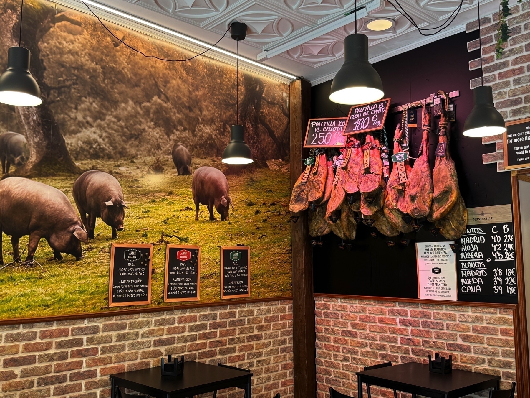 A cozy, rustic jamón shop corner with a photo mural of black Iberian pigs grazing in a wooded pasture, reinforcing the source of the high-quality meat. On the adjacent wall, several paletillas (front legs of jamón) hang beneath signs noting their type and price. Informational plaques explain the feeding and curing process, and a small table with chairs adds a welcoming touch to the tasting area.