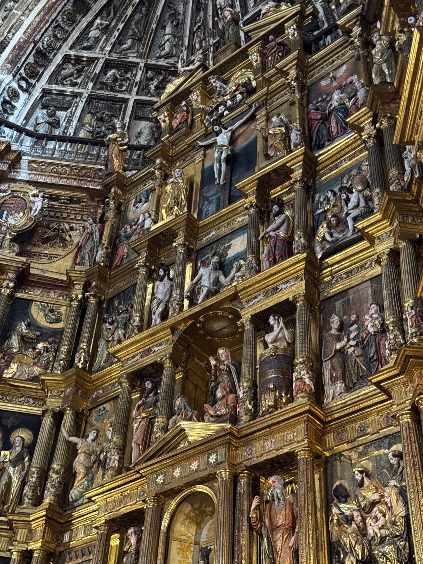Close-up of the upper portion of the altarpiece, highlighting expressive sculpted figures arranged in narrative scenes. The gold-trimmed architectural elements frame each panel, while painted reliefs and figures convey intense emotion and devotion.