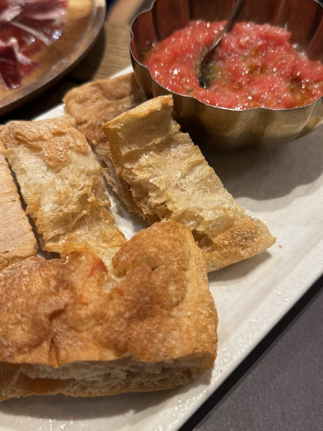 Crisp pieces of rustic bread served beside a brass bowl of grated tomato topping with a spoon, prepared in the style of pan con tomate.