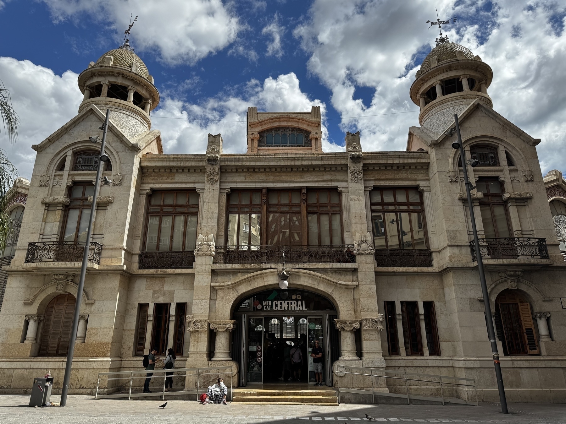 Front facade of Mercat Central de València, showcasing a symmetrical Art Nouveau design with two domed towers, arched entranceway, and decorative stonework under a partly cloudy sky.