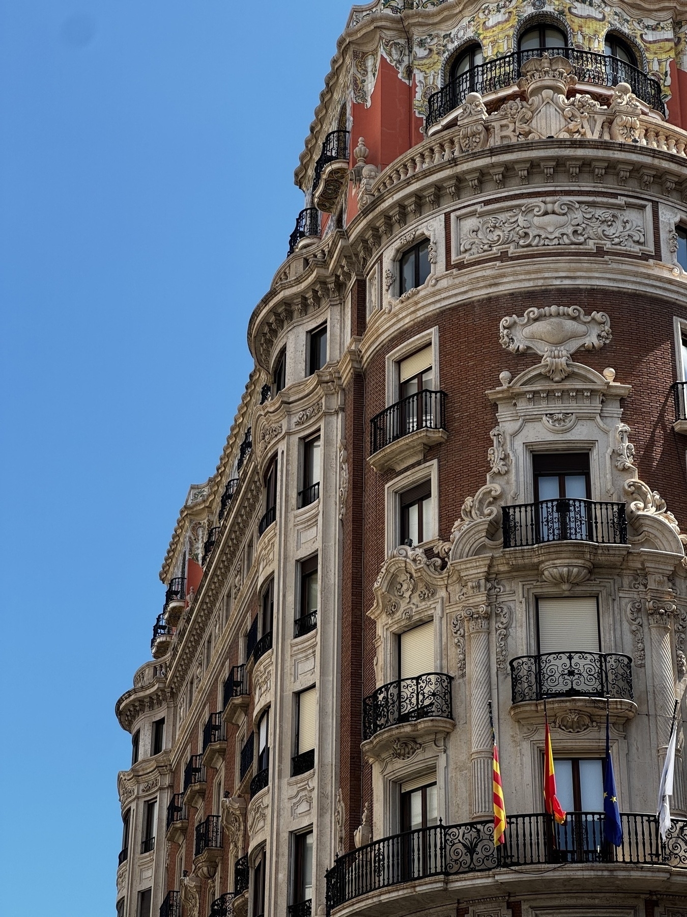 A grand corner building showcases ornate baroque architecture, with sculpted stonework, wrought iron balconies, and a striking red façade under a cloudless sky. Its intricate detailing and height convey elegance and history.