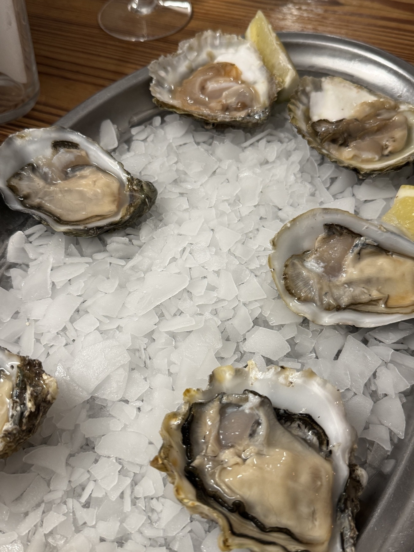 Six fresh oysters displayed in their half shells on a bed of crushed ice in a metal tray, garnished with two lemon wedges. A wine glass and part of a wooden table are visible in the background.