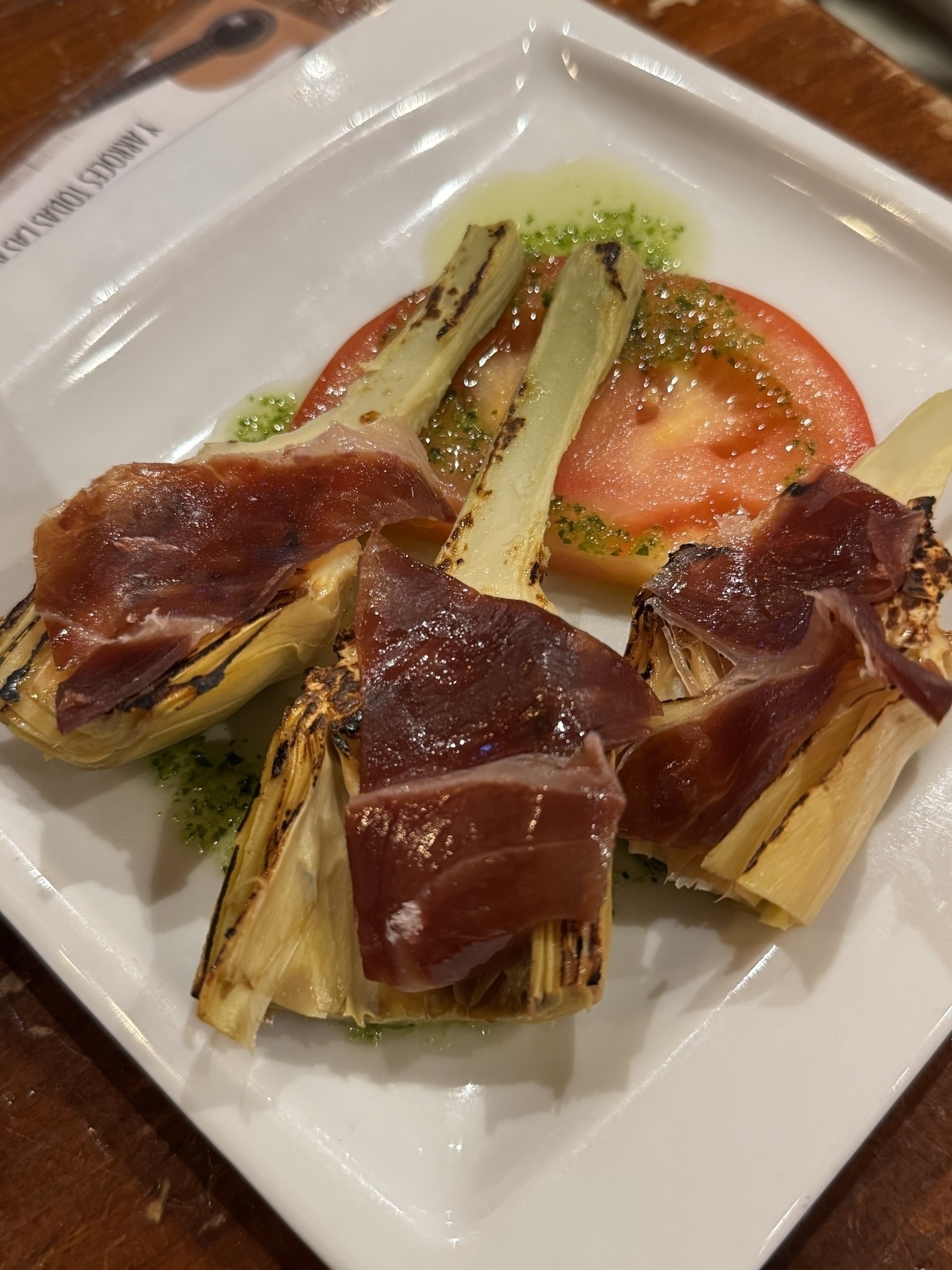 A square white plate with grilled artichoke quarters, slightly charred at the edges, topped with thin slices of jamón ibérico. A slice of fresh tomato sits beneath with a drizzle of green herb oil. The balance of smoky vegetables and cured ham makes it visually and texturally appealing.