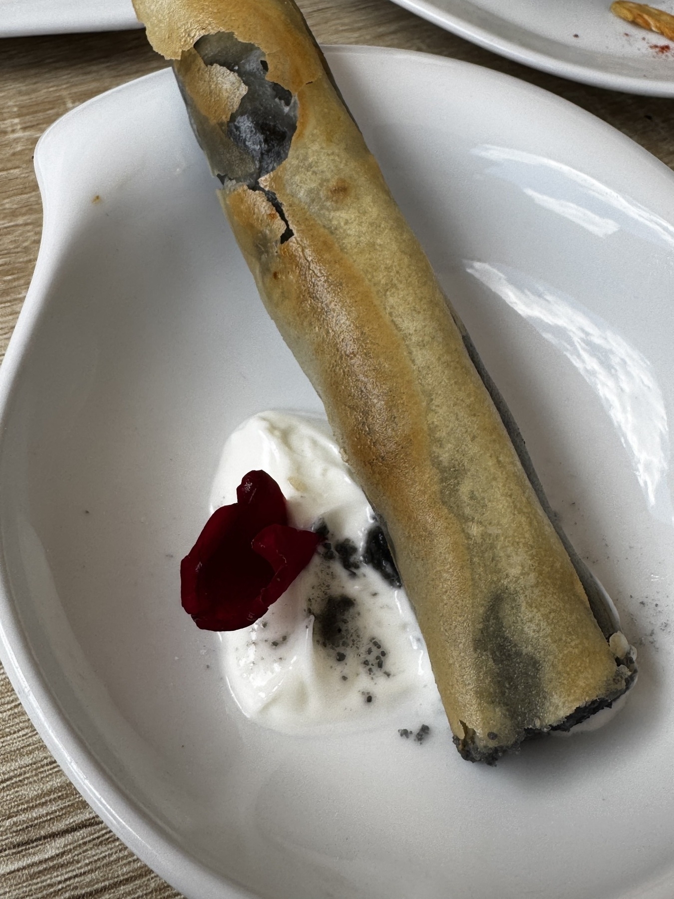 A crisp, golden pastry cigar filled with rich squid ink and seafood, artfully cracked to reveal its inky black interior, served over a dollop of smooth white sauce and garnished with a deep red flower petal for contrast.