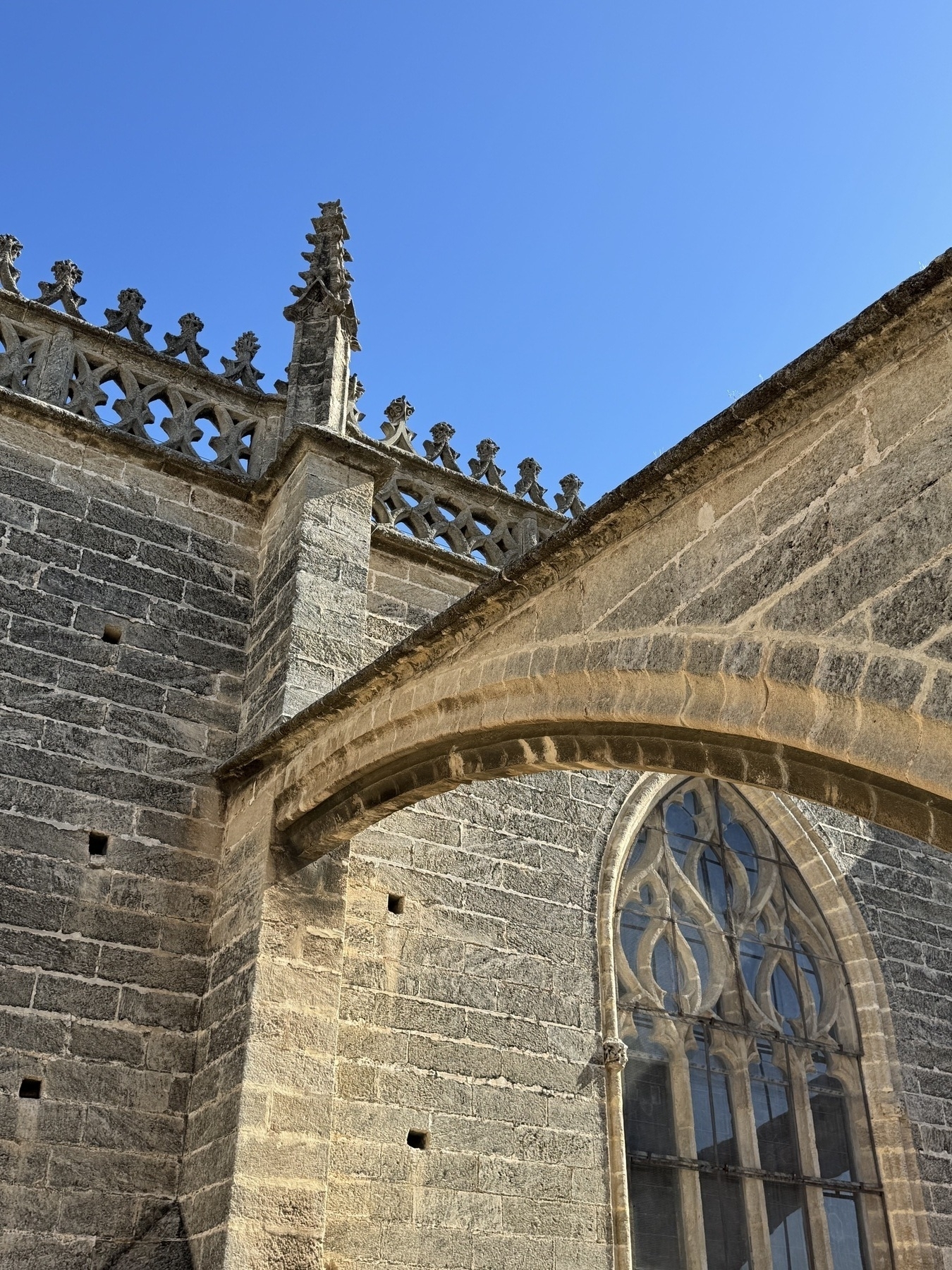 Close-up of a pointed arch window with elaborate stone tracery set into the cathedral’s grey stone wall, topped by decorative crenellations and small Gothic finials, under a vivid blue sky.