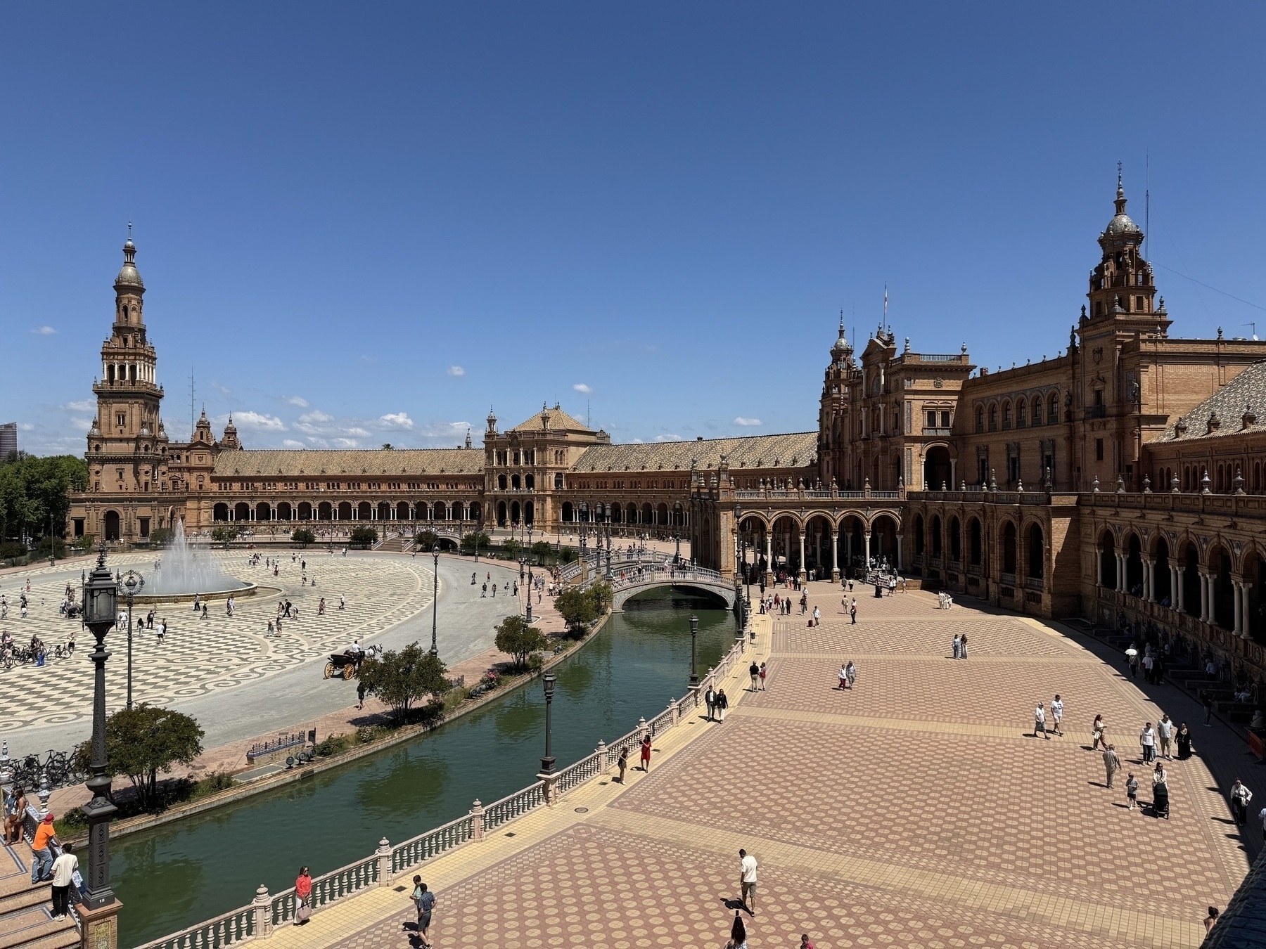A panoramic view of the Plaza de España reveals its sweeping semicircular shape, with richly detailed brick buildings, two towering spires, and arched arcades. A wide, patterned plaza with a large fountain and a curving canal is filled with people enjoying the clear day.