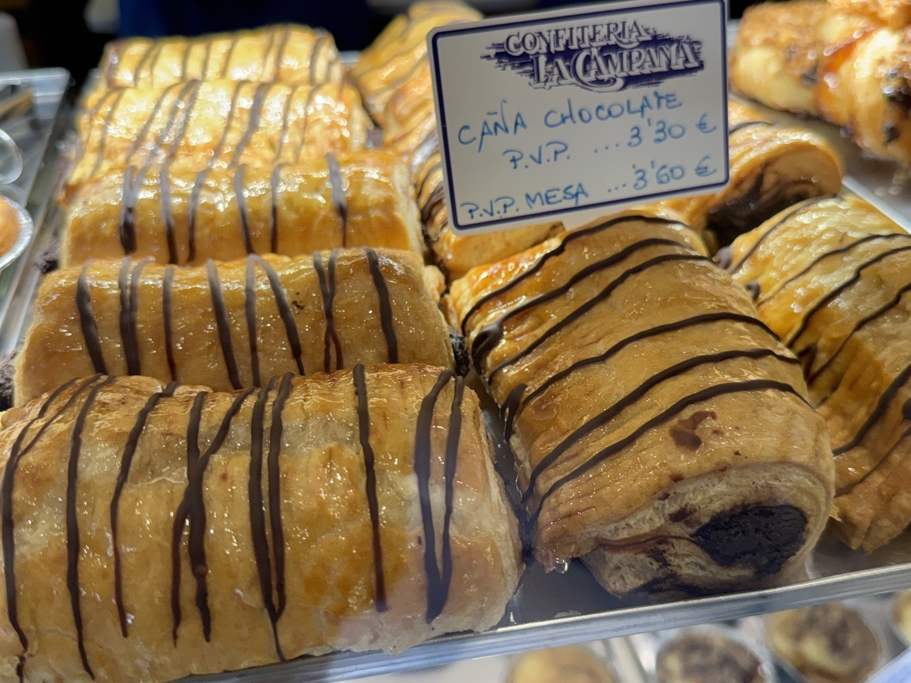 Pastry display at Confitería La Campana featuring “Caña Chocolate,” rectangular puff pastries with a shiny golden finish, drizzled generously with dark chocolate stripes. The pastries are arranged in neat rows, emphasizing their crisp, flaky layers and gooey chocolate filling, which makes them look rich, sweet, and irresistibly satisfying.